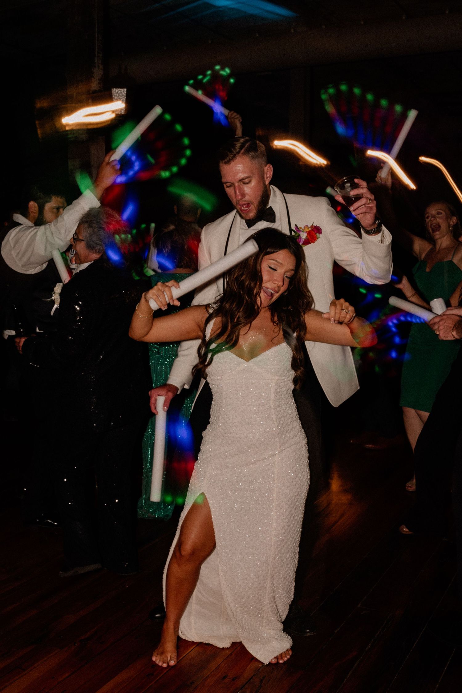 Bride and groom energetically dancing at a dark wedding reception hall with neon light props that create light trails
