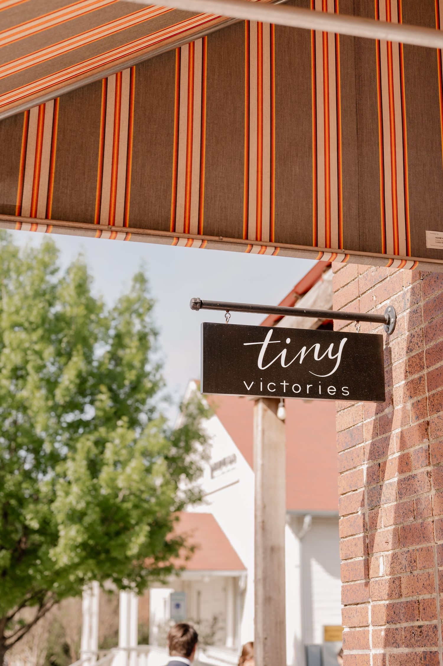 Striped awning with Tiny Victories sign mounted on brick wall.