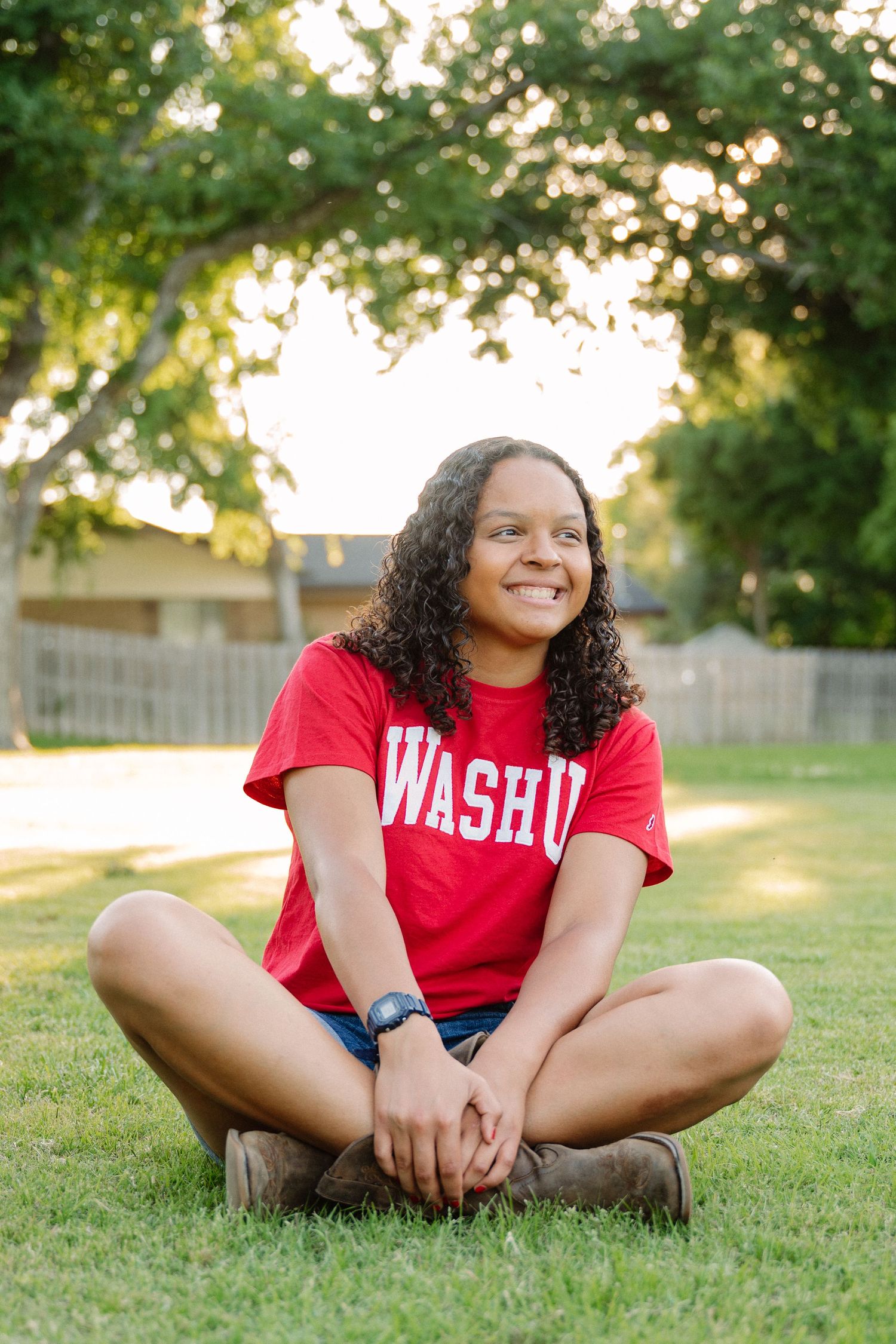 Senior sitting and smiling in her college tshirt and boots in a large park in Richardson, Texas for a senior photo session