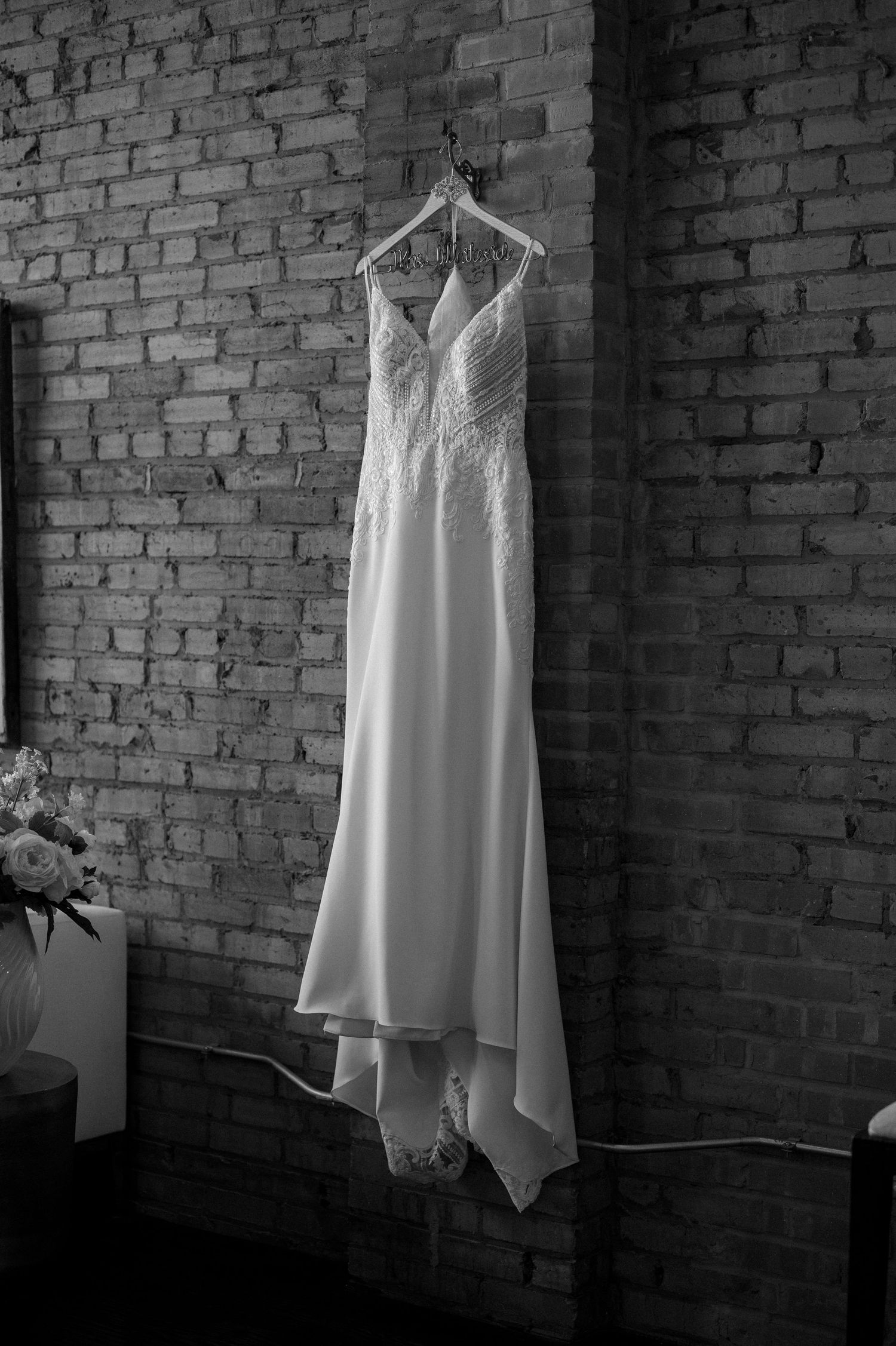 A white wedding dress hangs on display against a rustic brick wall in dramatic black and white lighting.