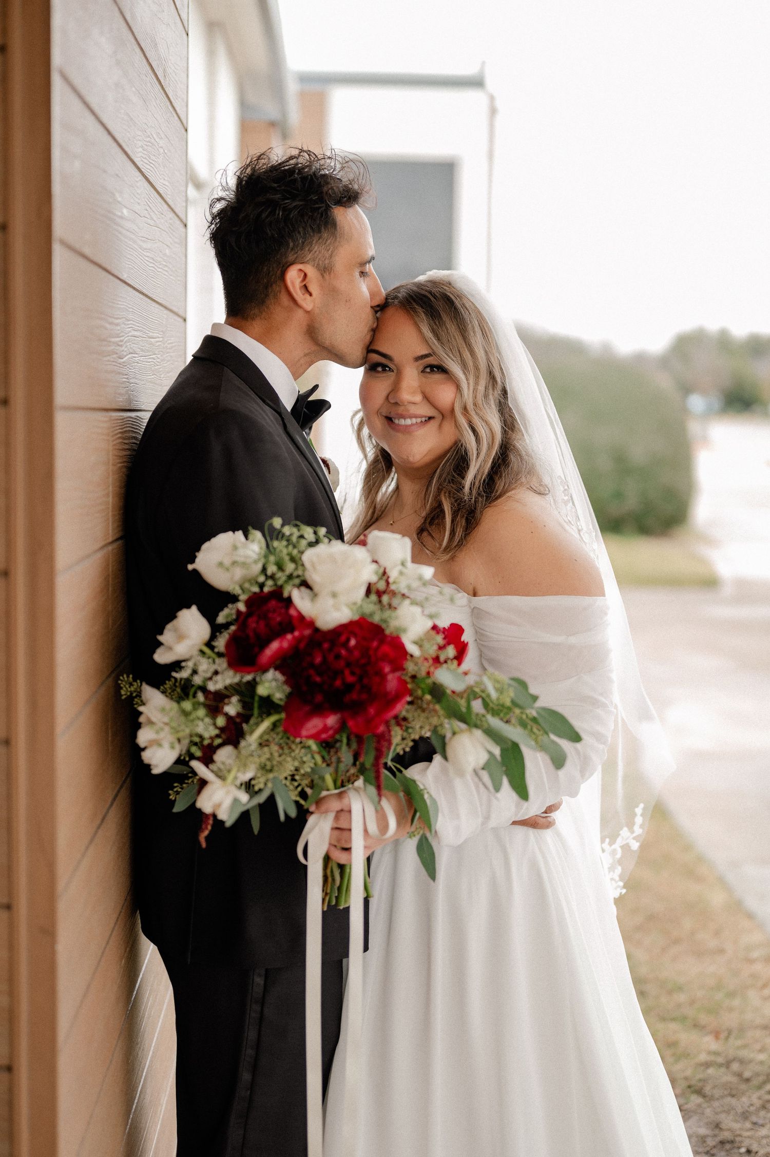 Bride and groom share an intimate wedding moment against a wooden wall while holding a vibrant red and white bouquet.