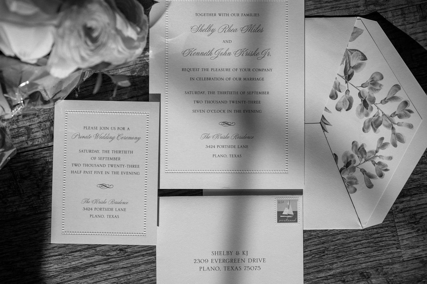 Wedding invitation suite featuring floral designs photographed in black and white on wooden surface.