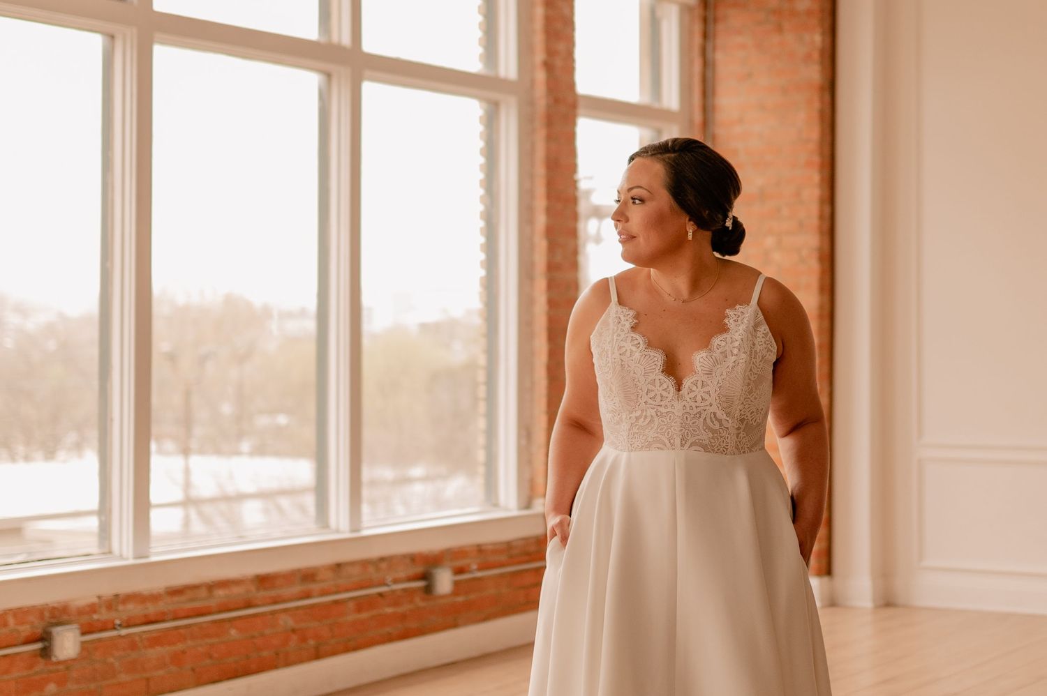 Bride in lace-detailed wedding dress gazes out large windows in elegant industrial venue.
