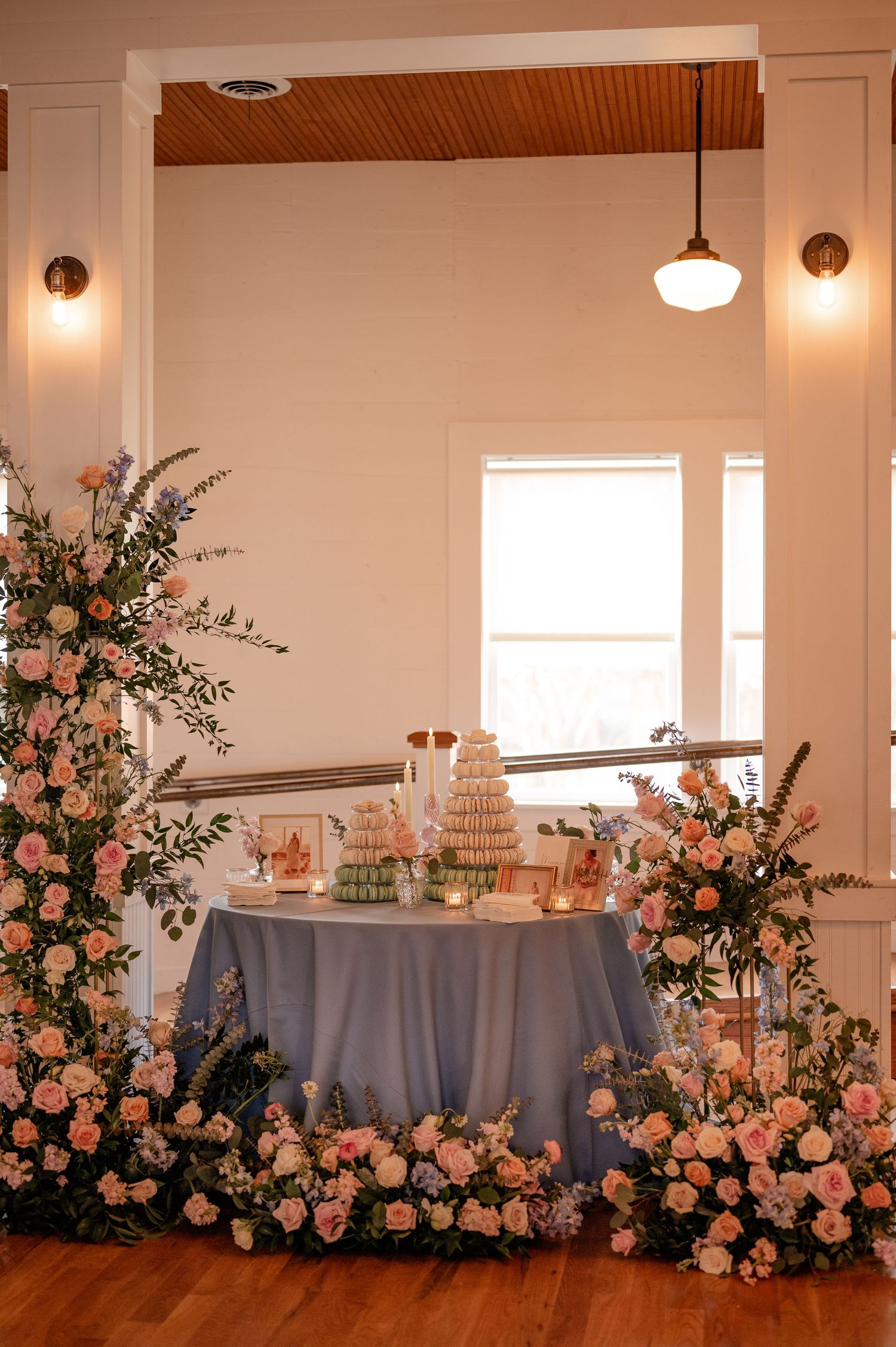 Wedding cake display table surrounded by lush pink floral arrangements and white walls with pendant lighting.