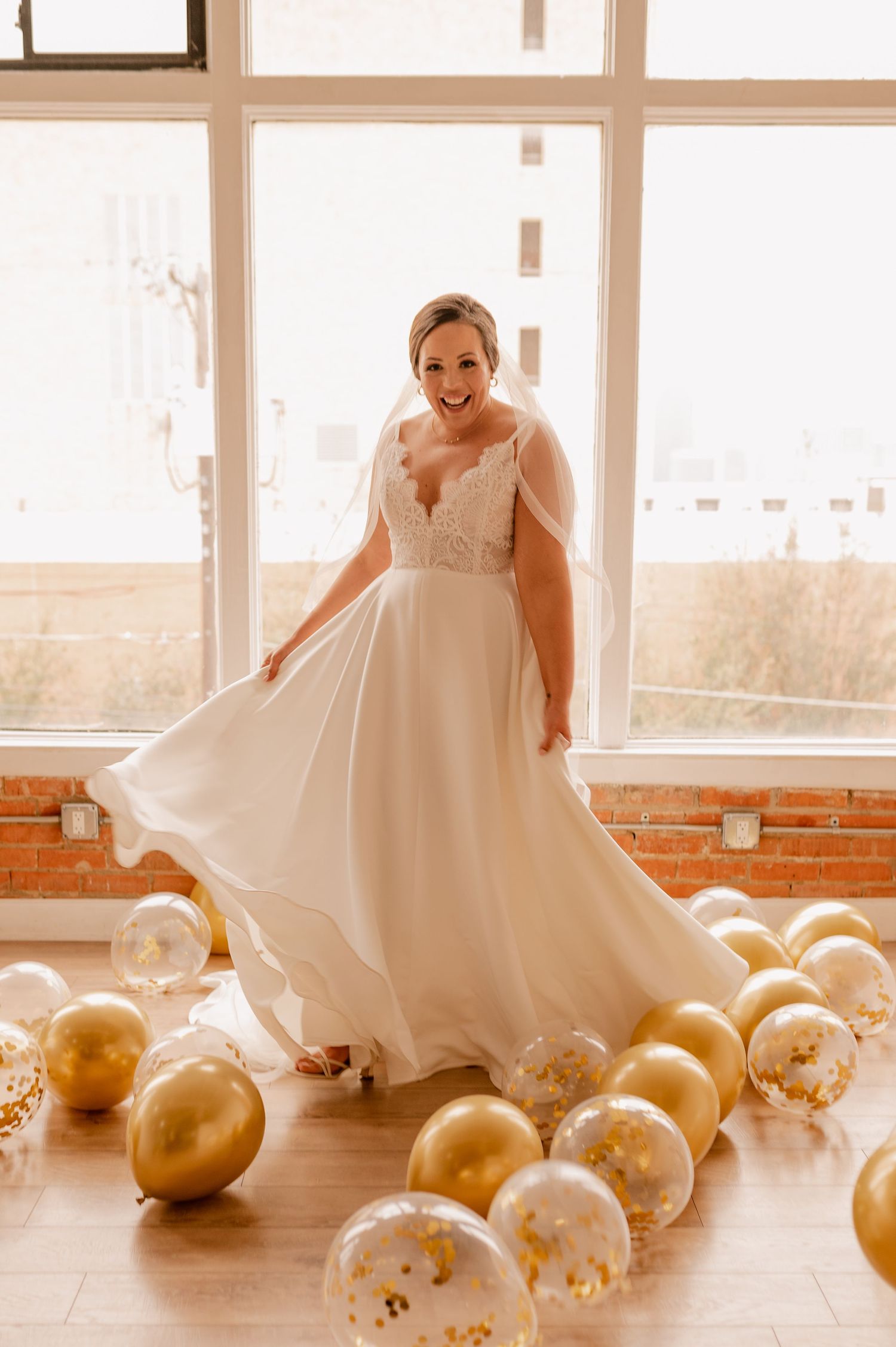 Bride twirling in white gown surrounded by gold balloons in sunlit industrial space.
