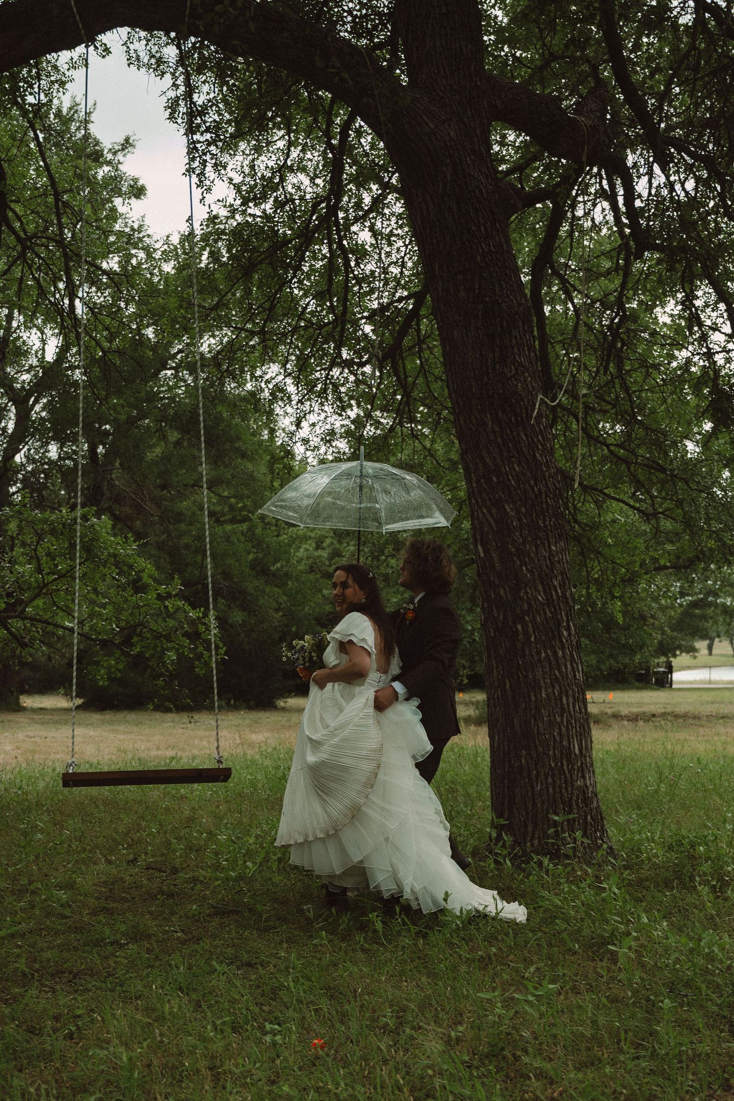 Moody candid photo of wedding couple walking through a large tree under an umbrella on their rainy wedding day in Ft. Worth, TX