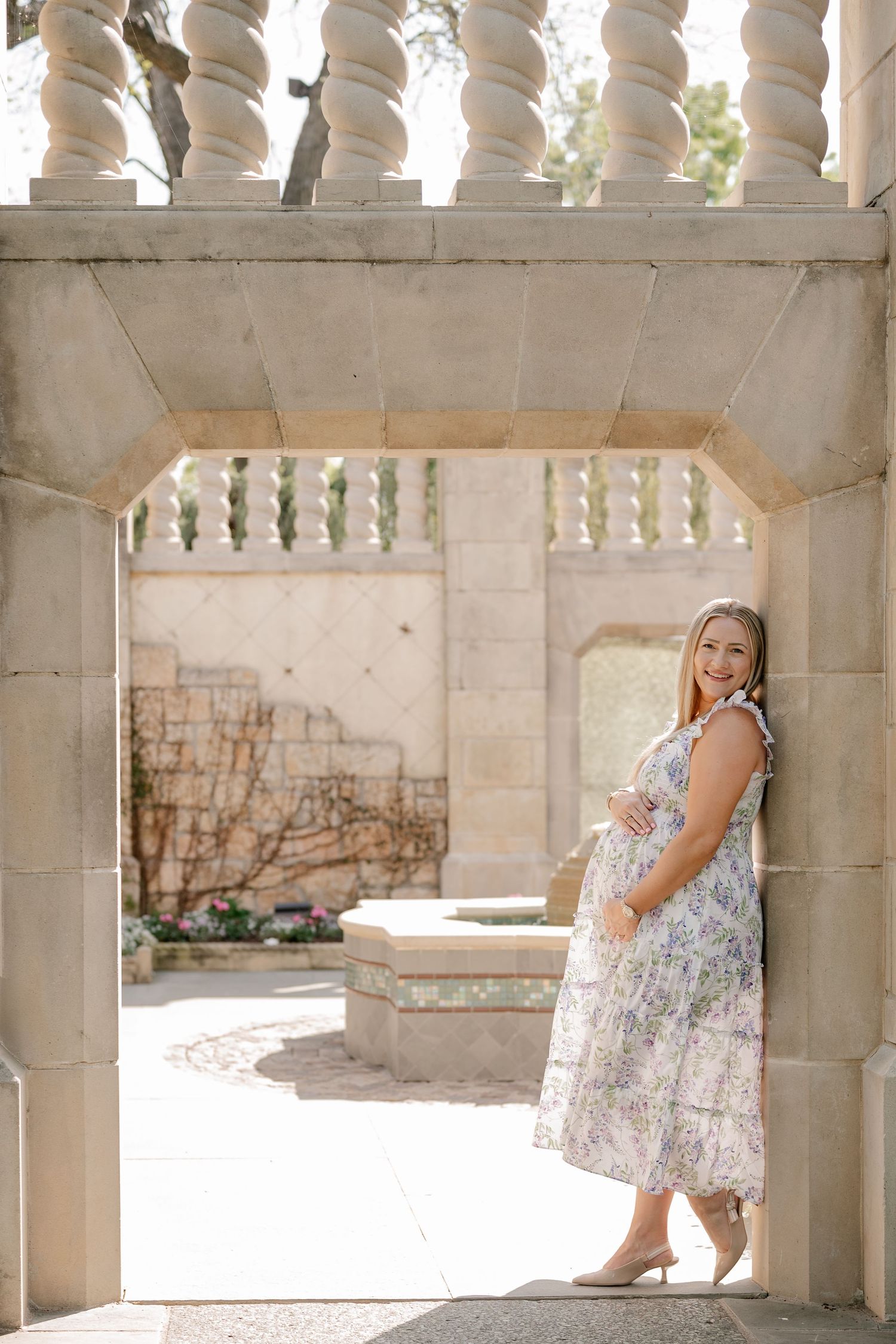 Woman leaning up against a stone archway holding her pregnant belly for a Spring maternity session at the Dallas Arboretum