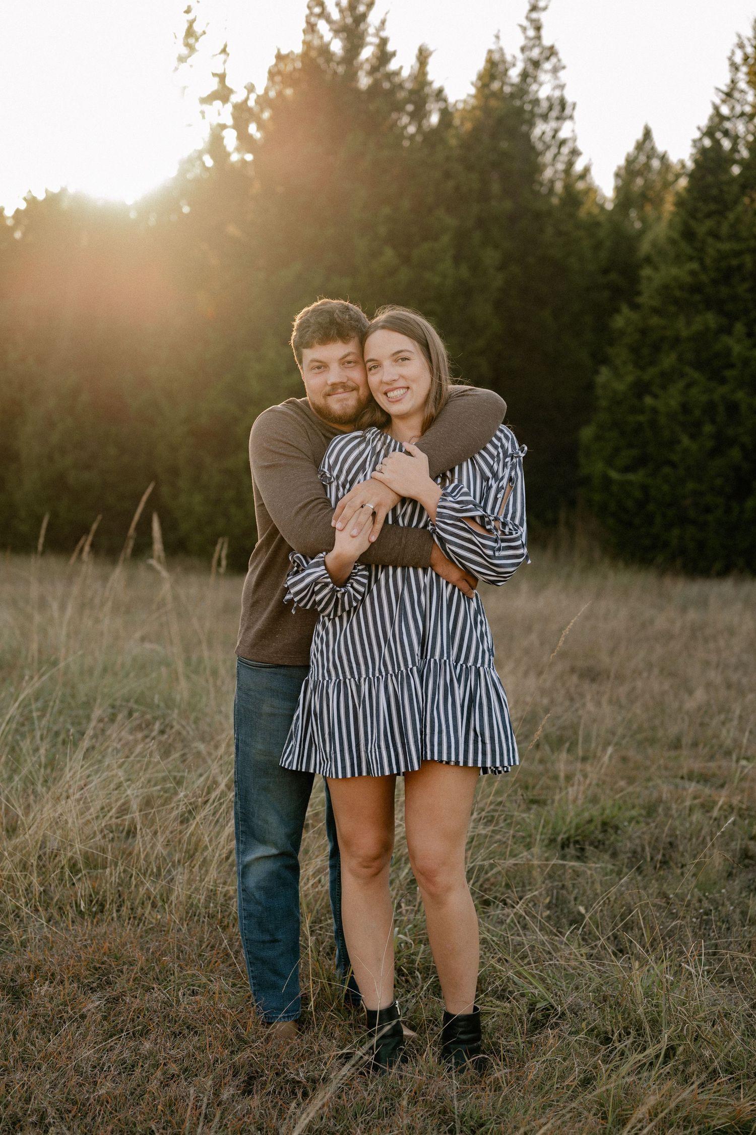 Couple standing and hugging in grassy autumn field with treelined backdrop during engagement photo session.