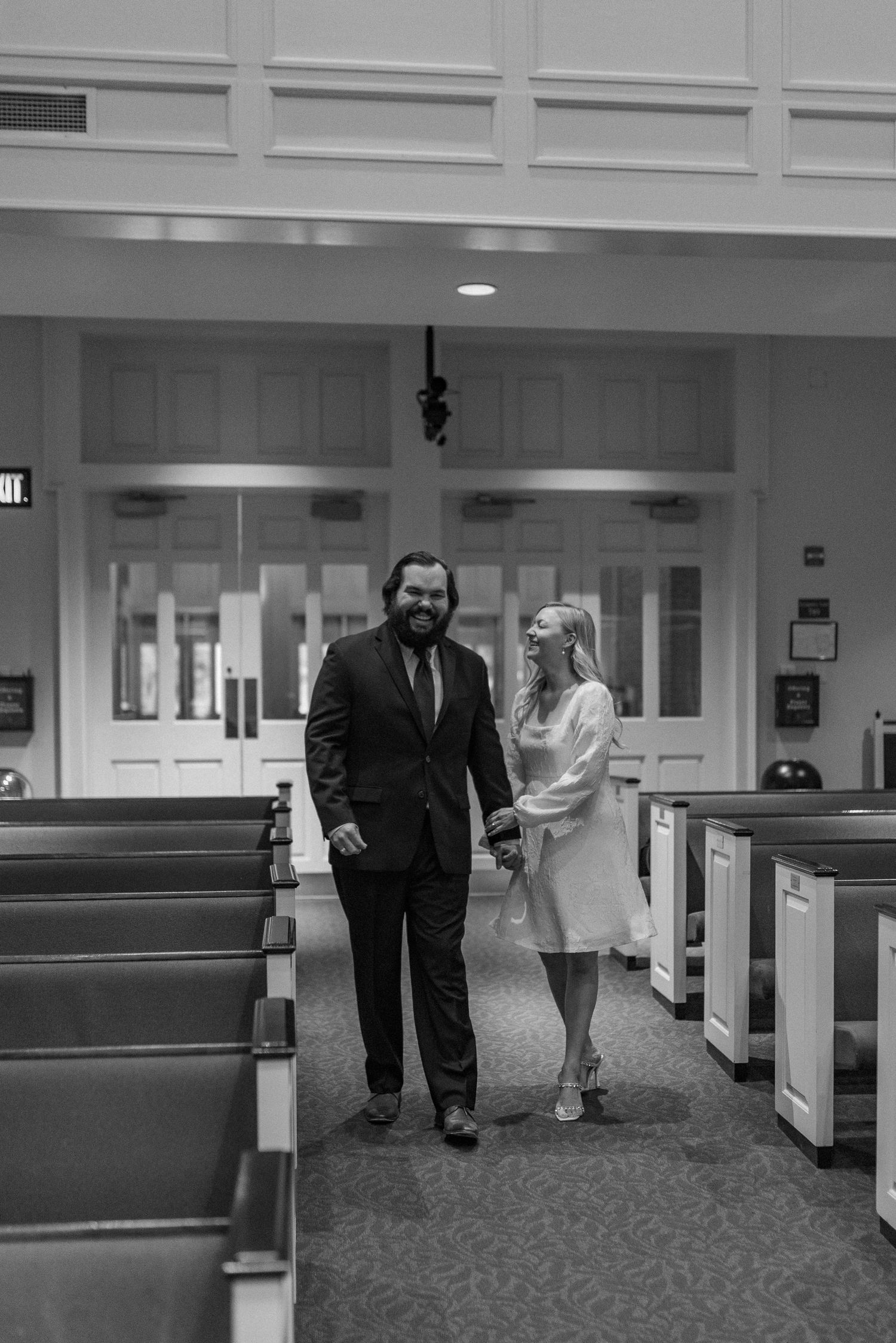 Black and white photo of people walking down a carpeted hallway in formal attire.