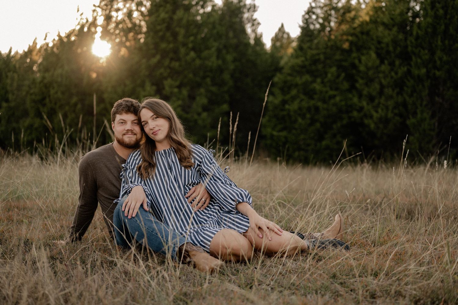 Married couple cuddling while sitting during a casual fall couple session in a large tree lined field in during golden hour
