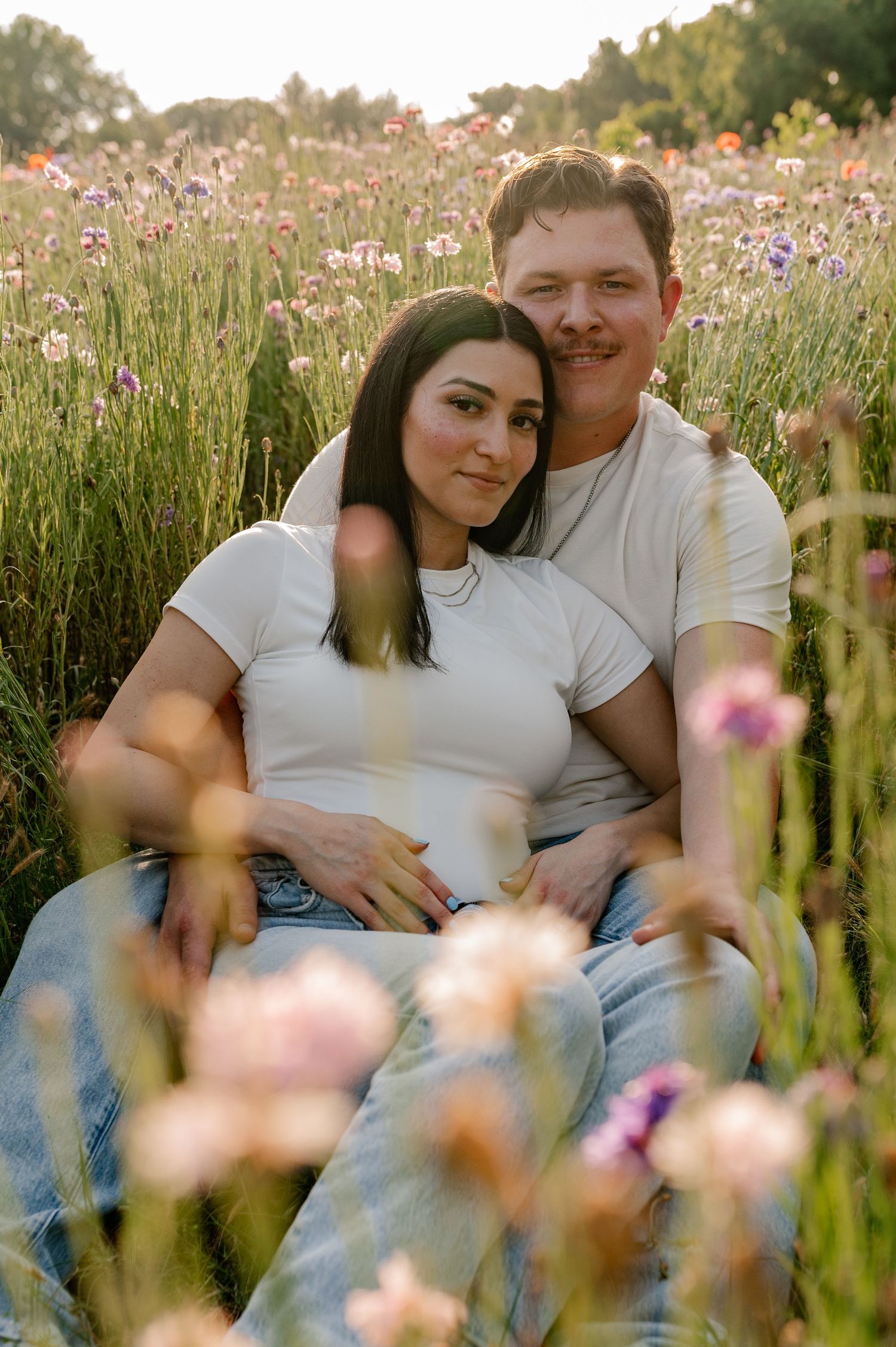 Couple sitting in tall pink wildflowers in bright light for a Spring maternity announcement during a couple photo session in Richardson, TX