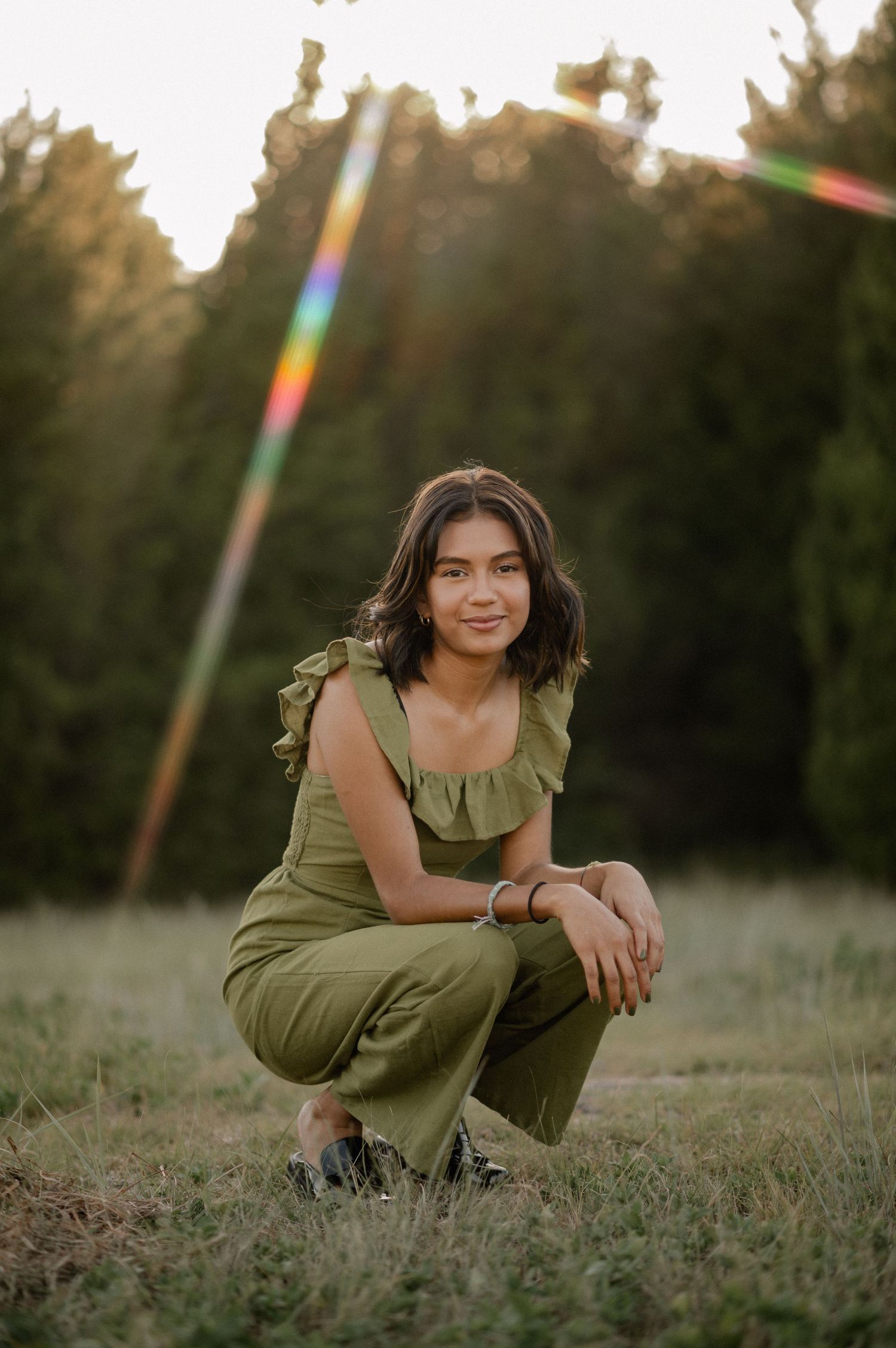 Girl in a green jumpsuit in a large field with a rainbow sun leak squatted down posing for a summer portrait session in Sunnyvale, Texas
