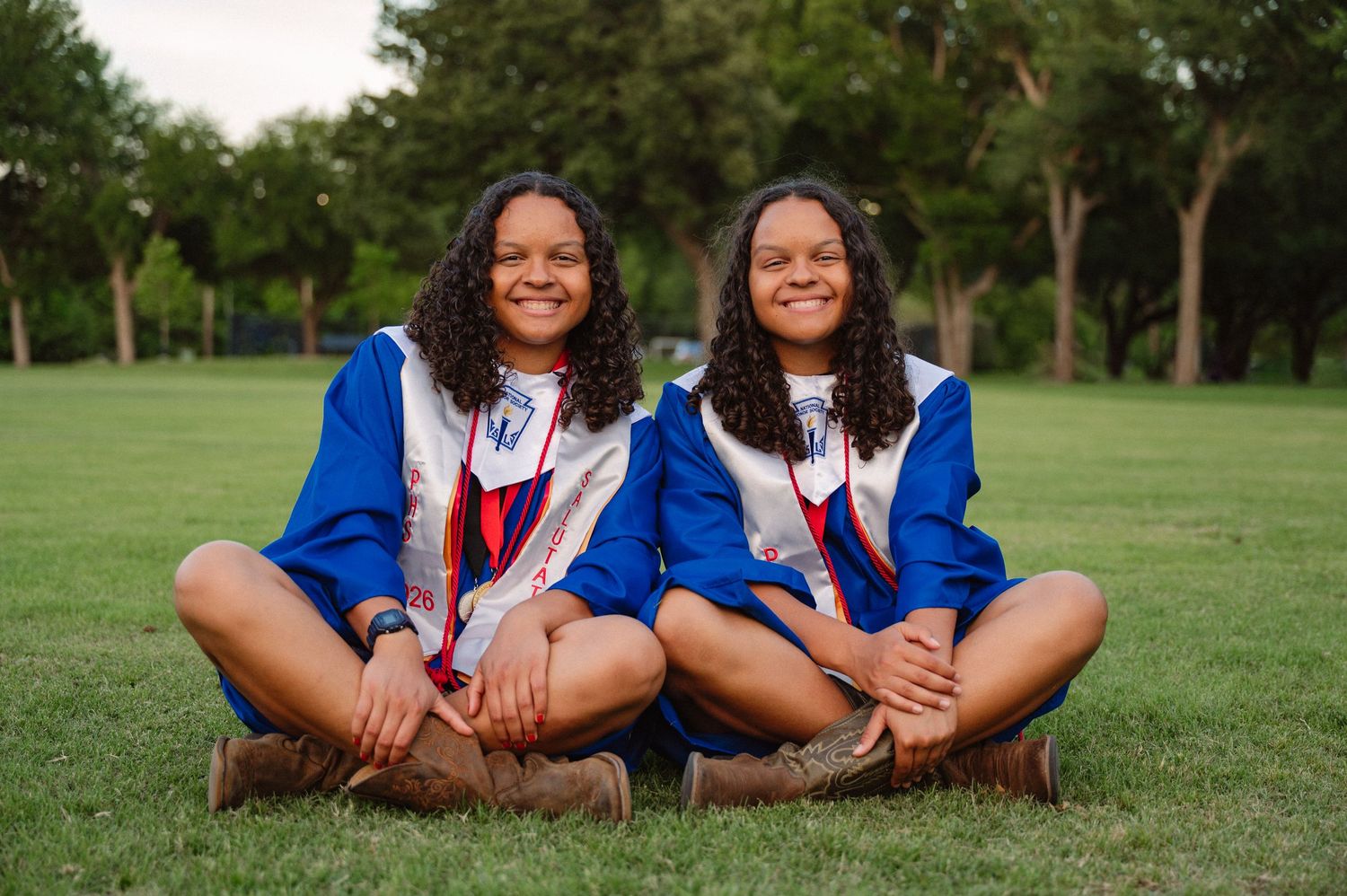 Senior twins in their grad gowns sitting side by side in a large tree-filled park in Richardson, Texas for a senior photo session