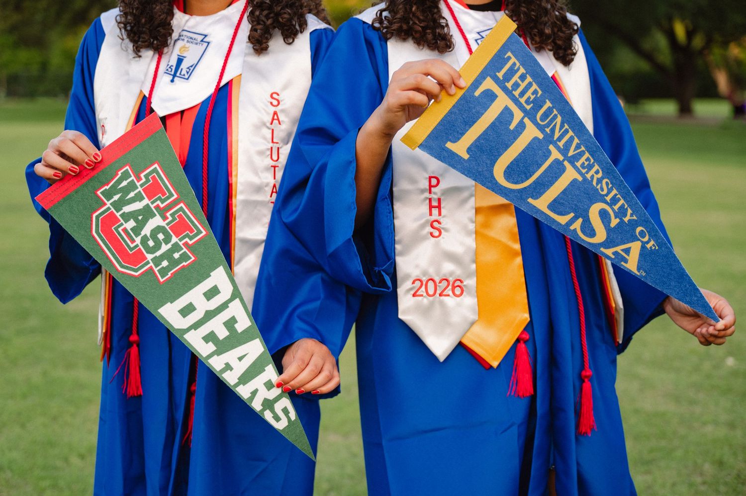 Senior twins in their grad gowns holding their college pennants in a large tree-filled park in Richardson, Texas for a senior photo session