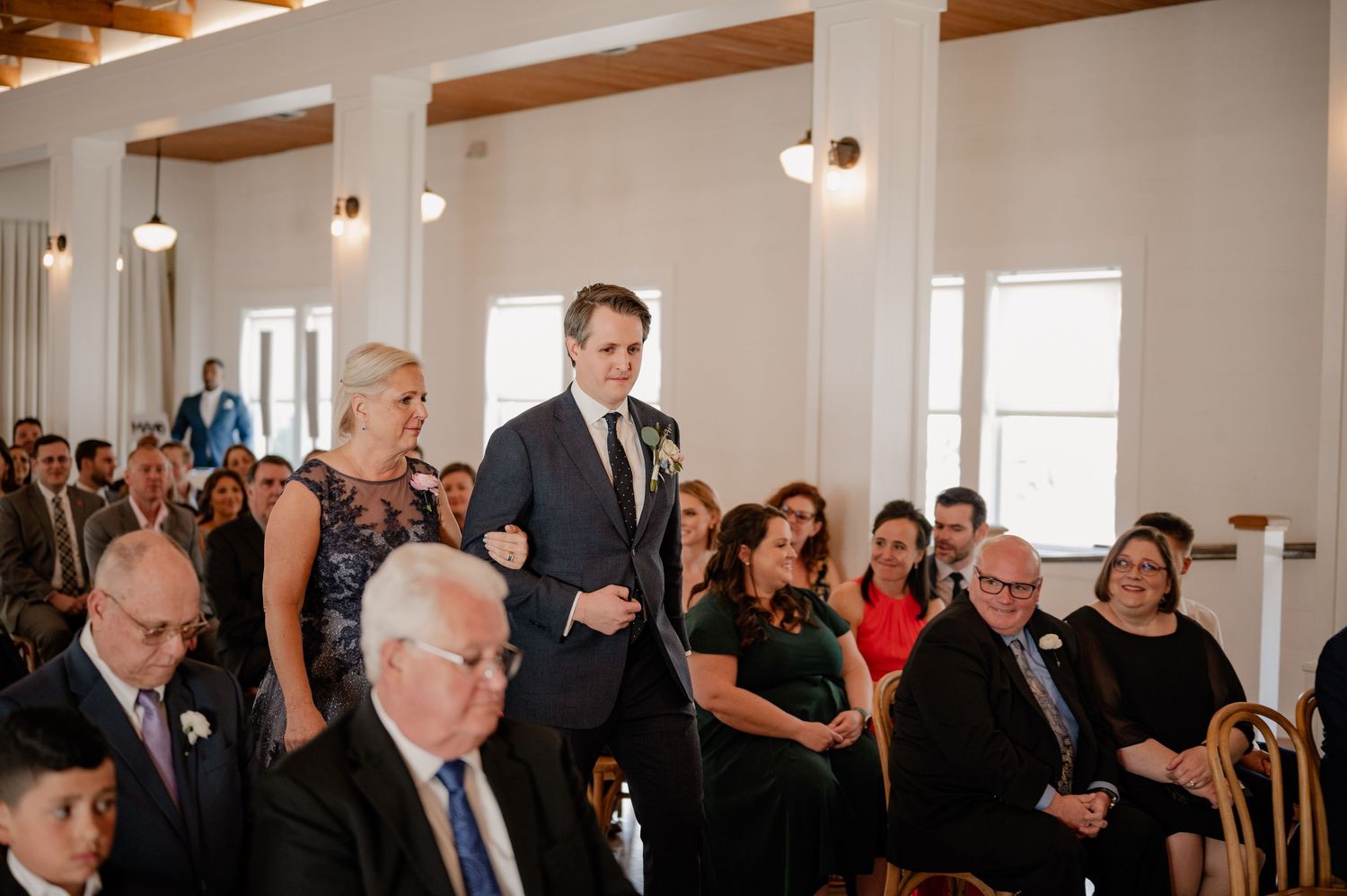 Wedding ceremony guests seated in a bright venue while groom and mother of the groom walk down the aisle