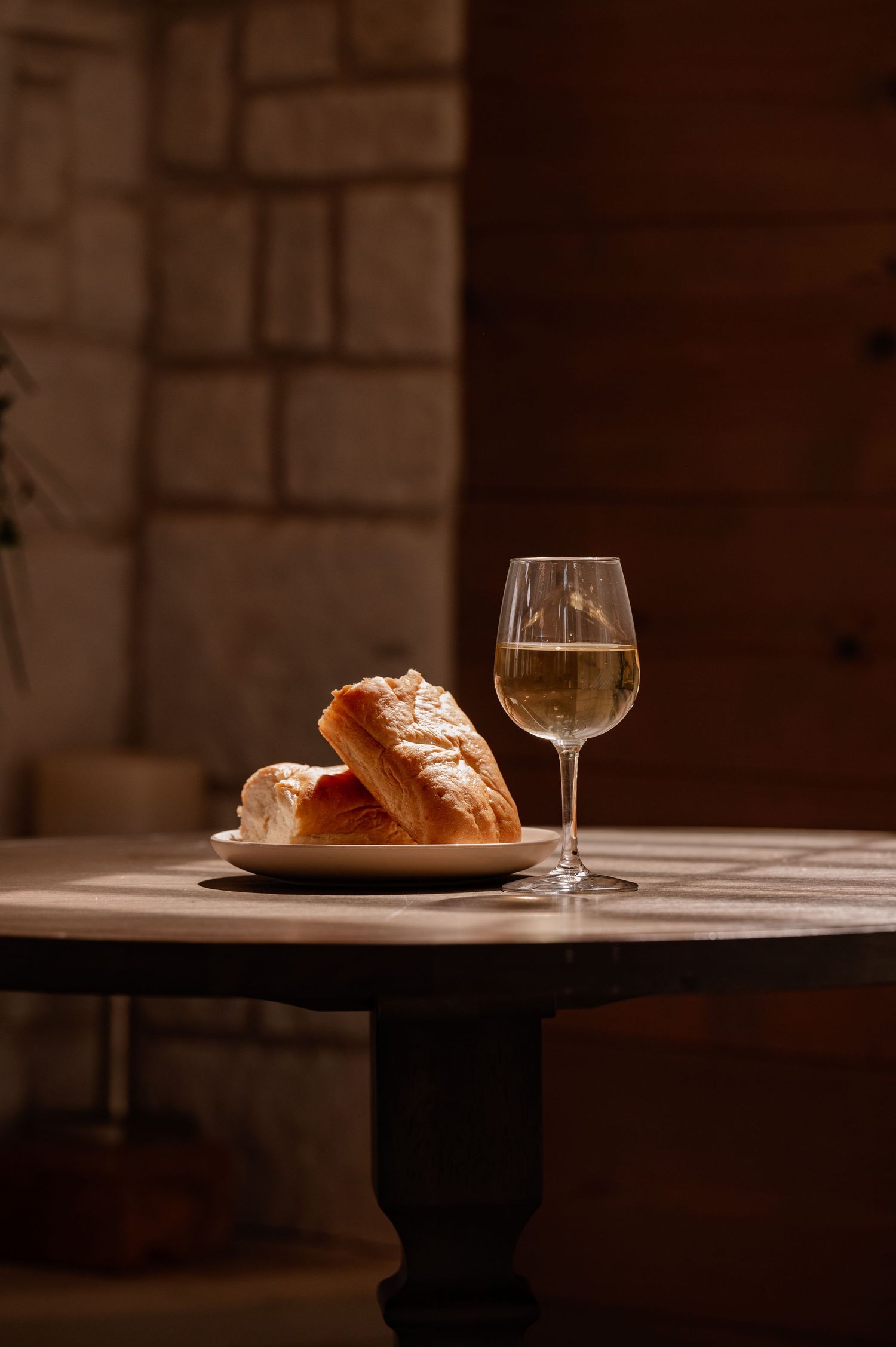 A glass of communion wine beside fresh bread on a plate, set on a rustic wooden table in warm, moody lighting.