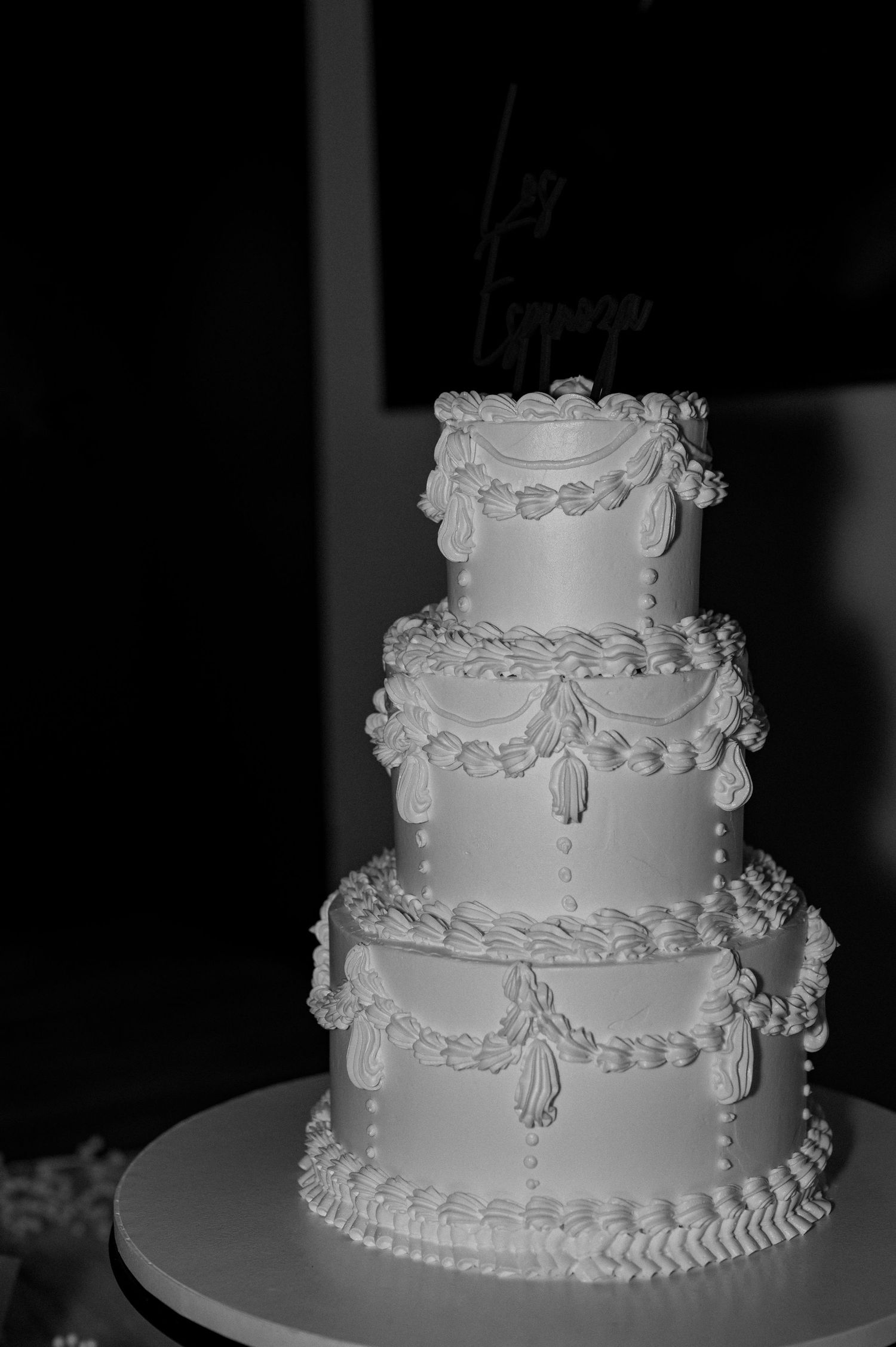 Elegant three-tier white wedding cake with intricate piped frosting details and decorative topper, captured in black and white.
