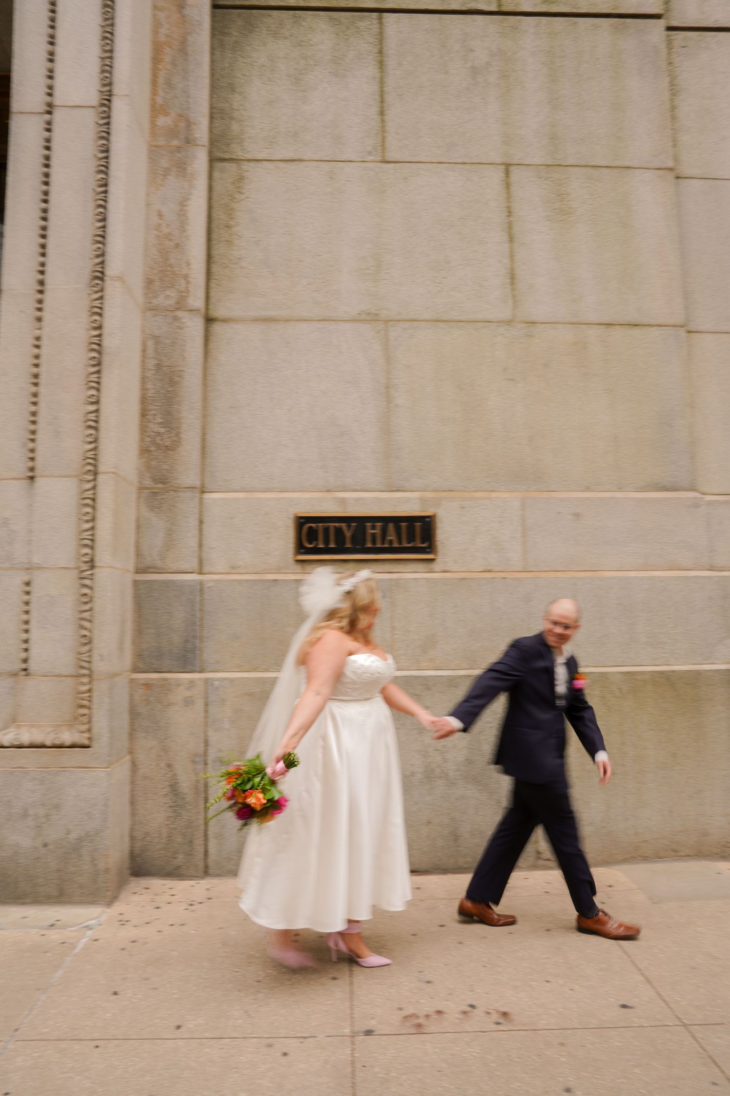Bride and groom holding hands walking past City Hall building on their wedding day.