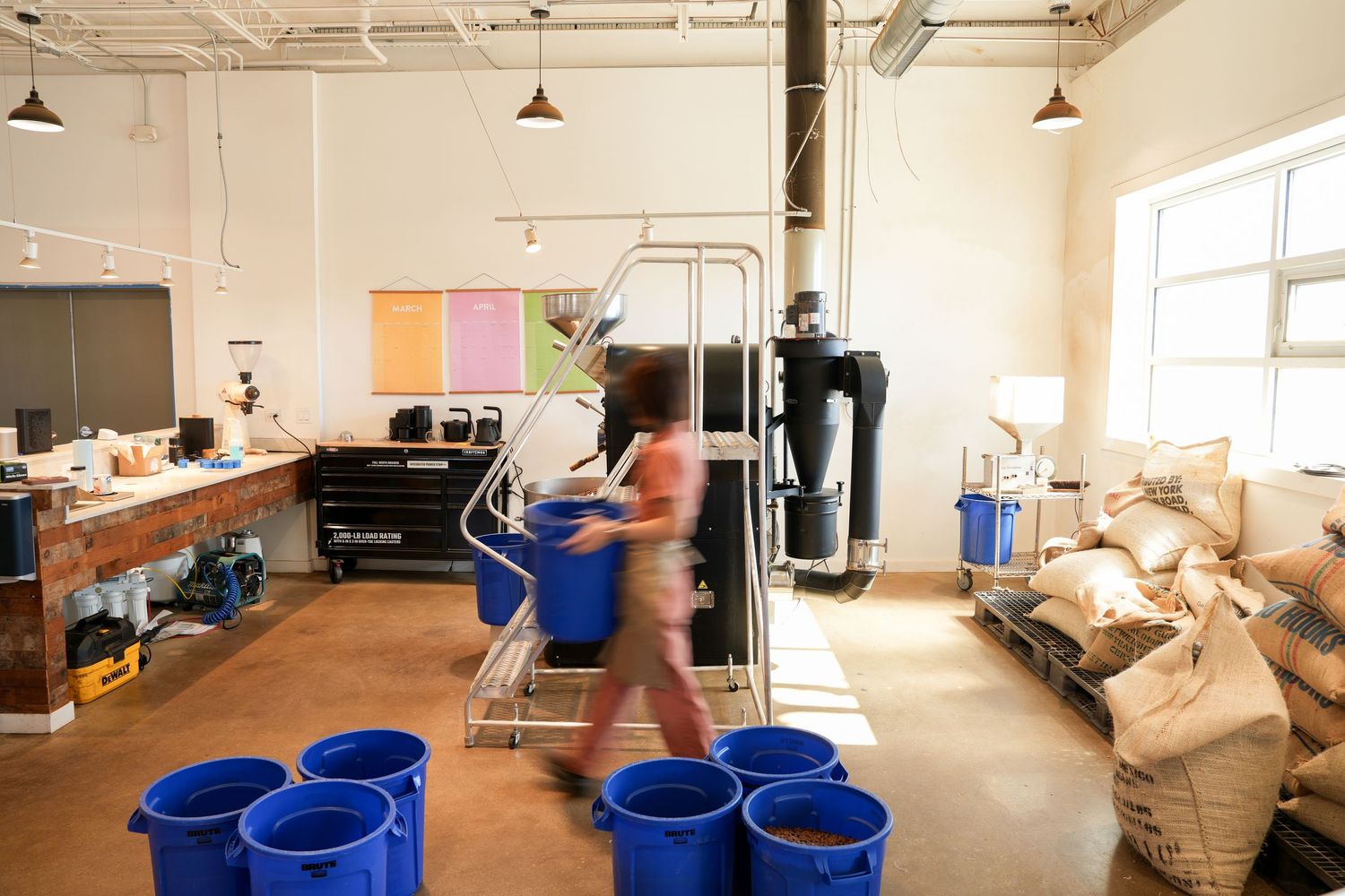 Industrial workspace with blue barrels, metal shelving, and burlap sacks, as a person walks through the busy facility.