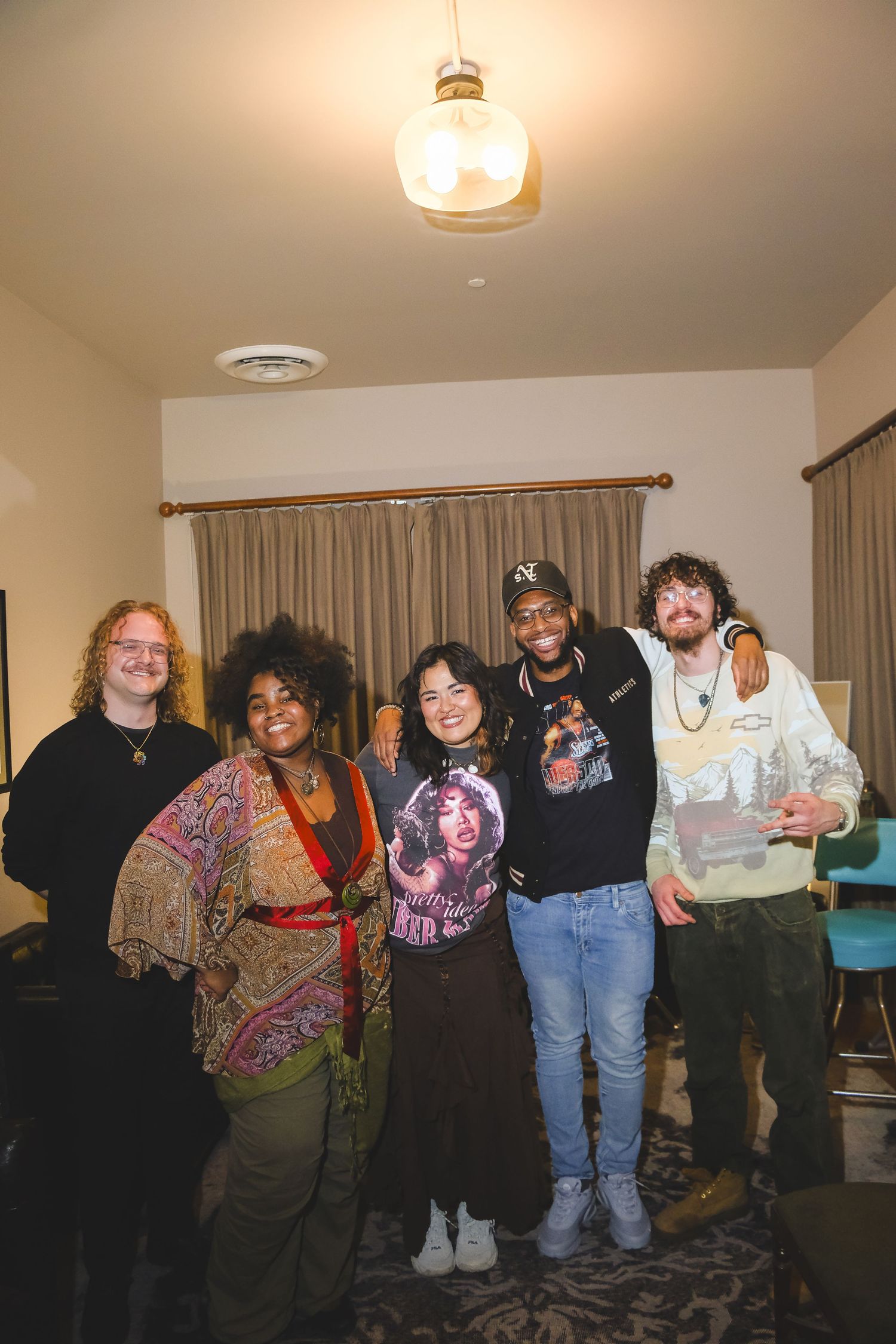 Five friends smiling together indoors under warm pendant light, dressed in casual and bohemian styles.