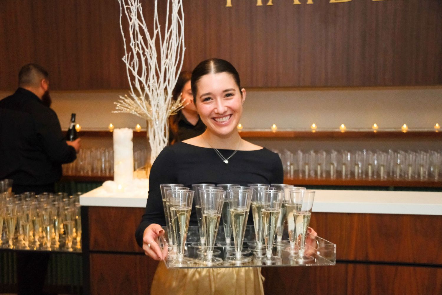 Rows of empty wine glasses lined up on a wooden bar counter with warm lighting in the background.