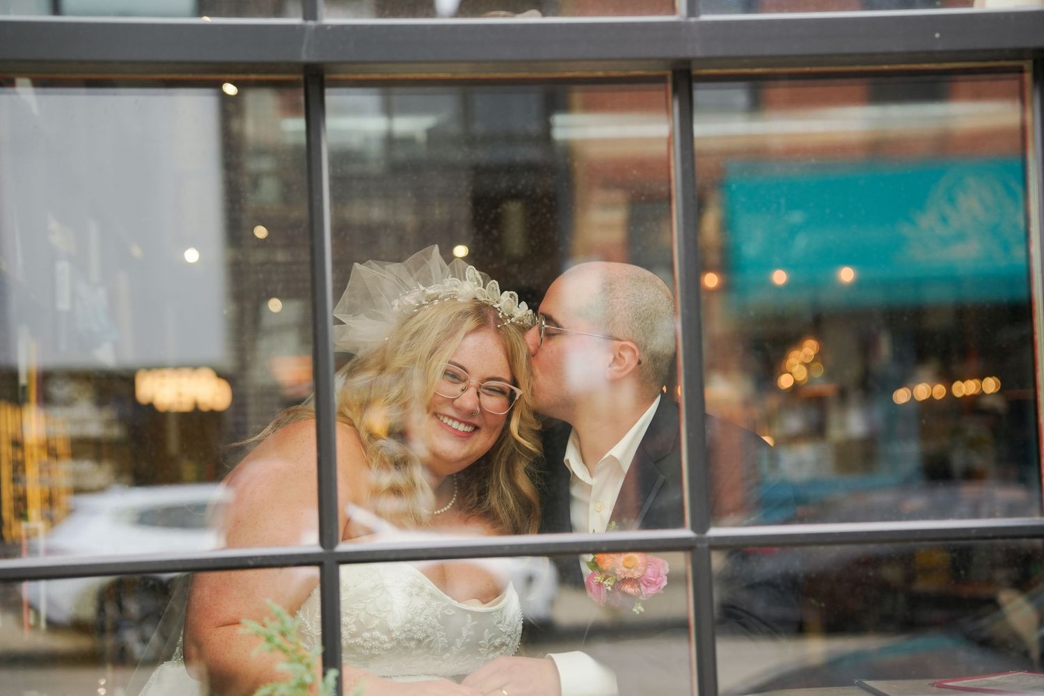 Bride and groom share a romantic kiss through a window, bride wearing a veil and lace dress on their wedding day.