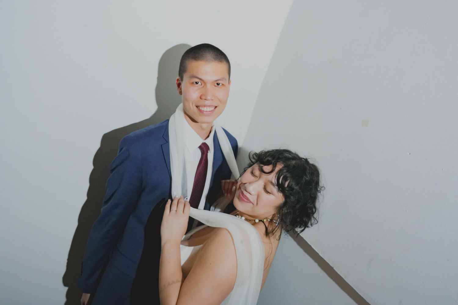 A smiling couple in wedding attire poses against a white wall, the groom in a navy suit and the bride in a white dress.