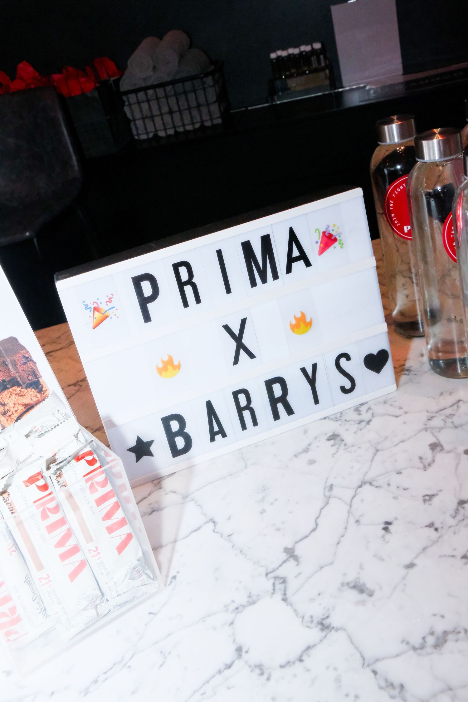 Light box sign reading Prima X Barrys with fire and party emojis displayed on a marble counter at a collaborative event.