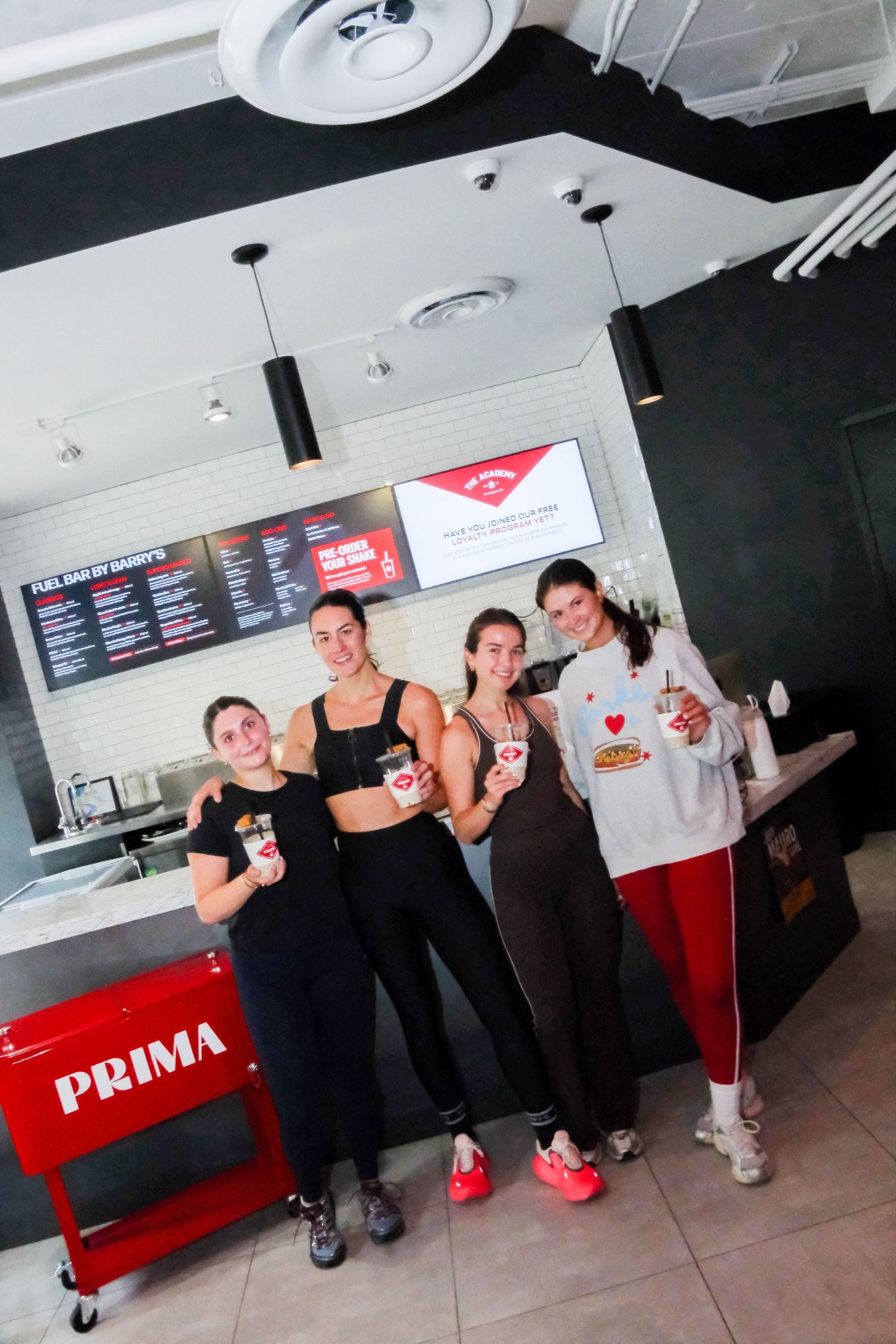 Four women posing together inside a Prima fitness cafe, holding food items near a red Prima cart.