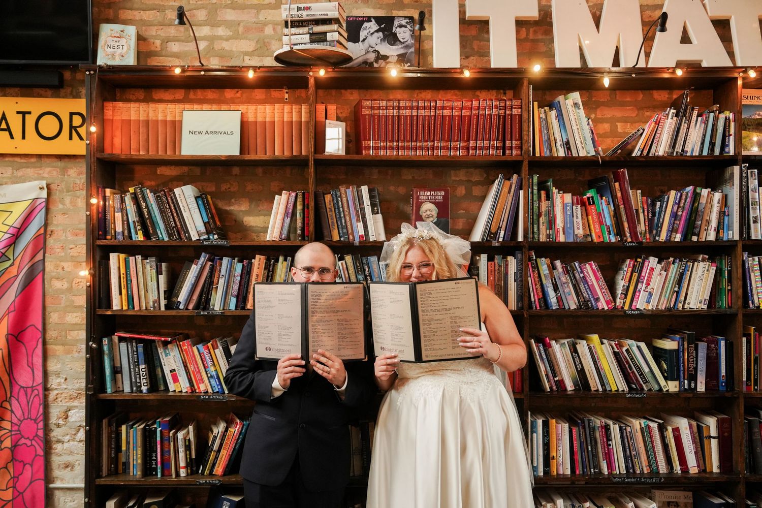 Couple in wedding attire hiding faces behind books in front of floor-to-ceiling bookshelves in a cozy bookstore.