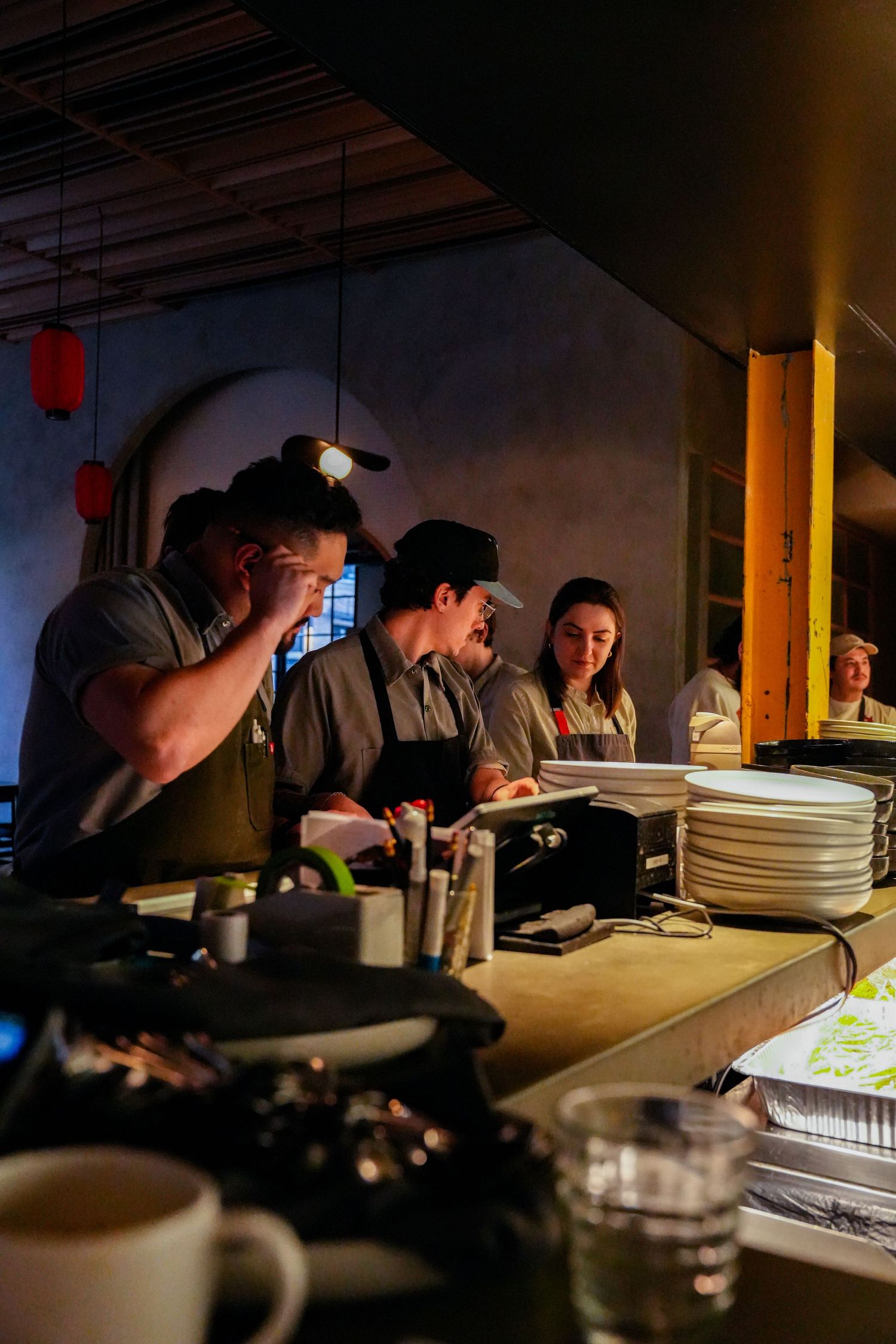 Restaurant kitchen staff working at a dimly lit counter with stacked plates and equipment during service.