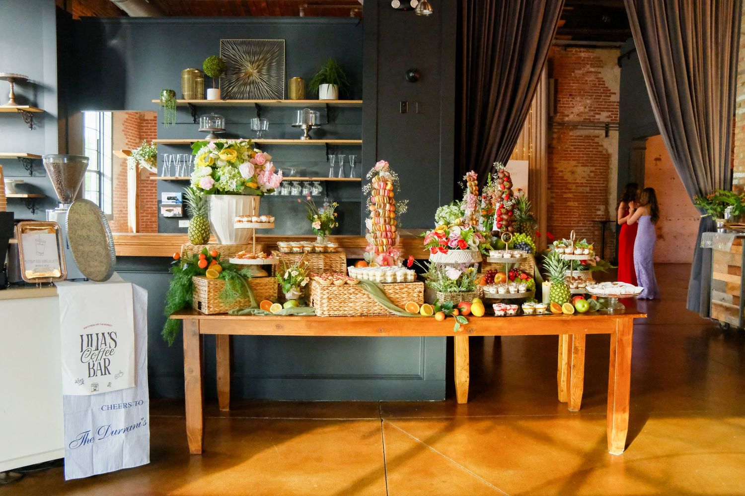 Elegant dessert bar table with colorful treats, greenery, and floral arrangements in a stylish event venue.