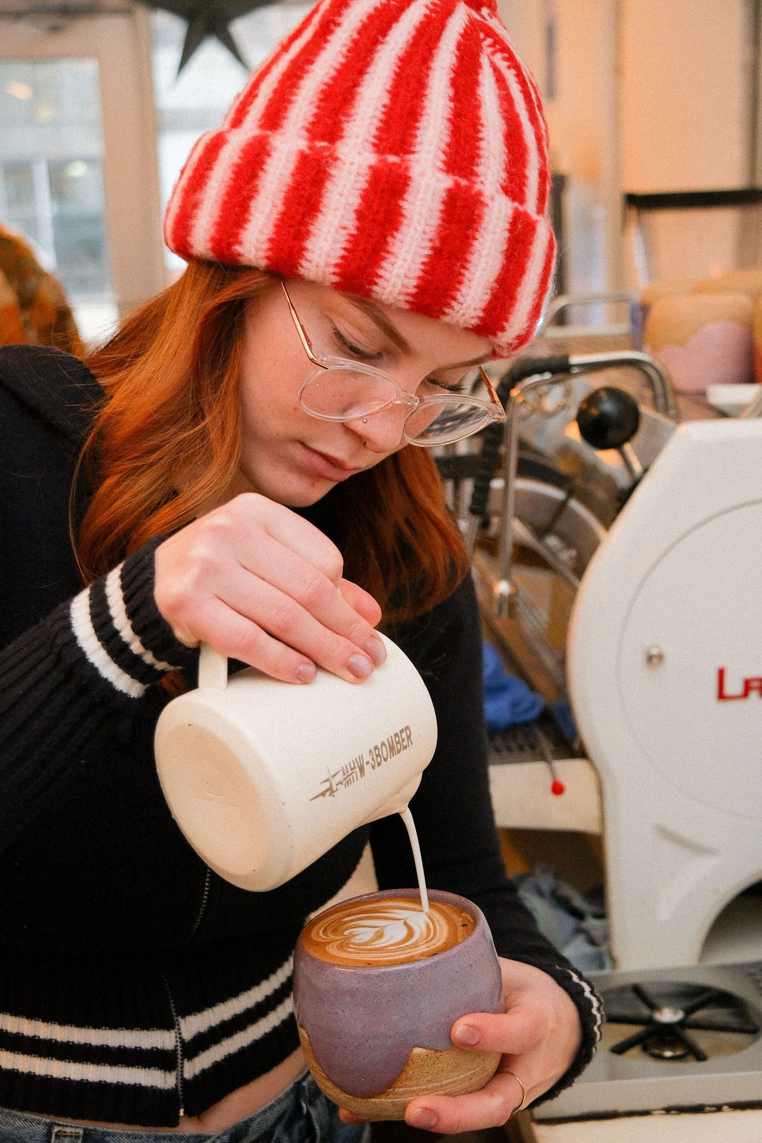 A barista in a red striped beanie carefully pours steamed milk into a latte, creating artistic coffee designs.