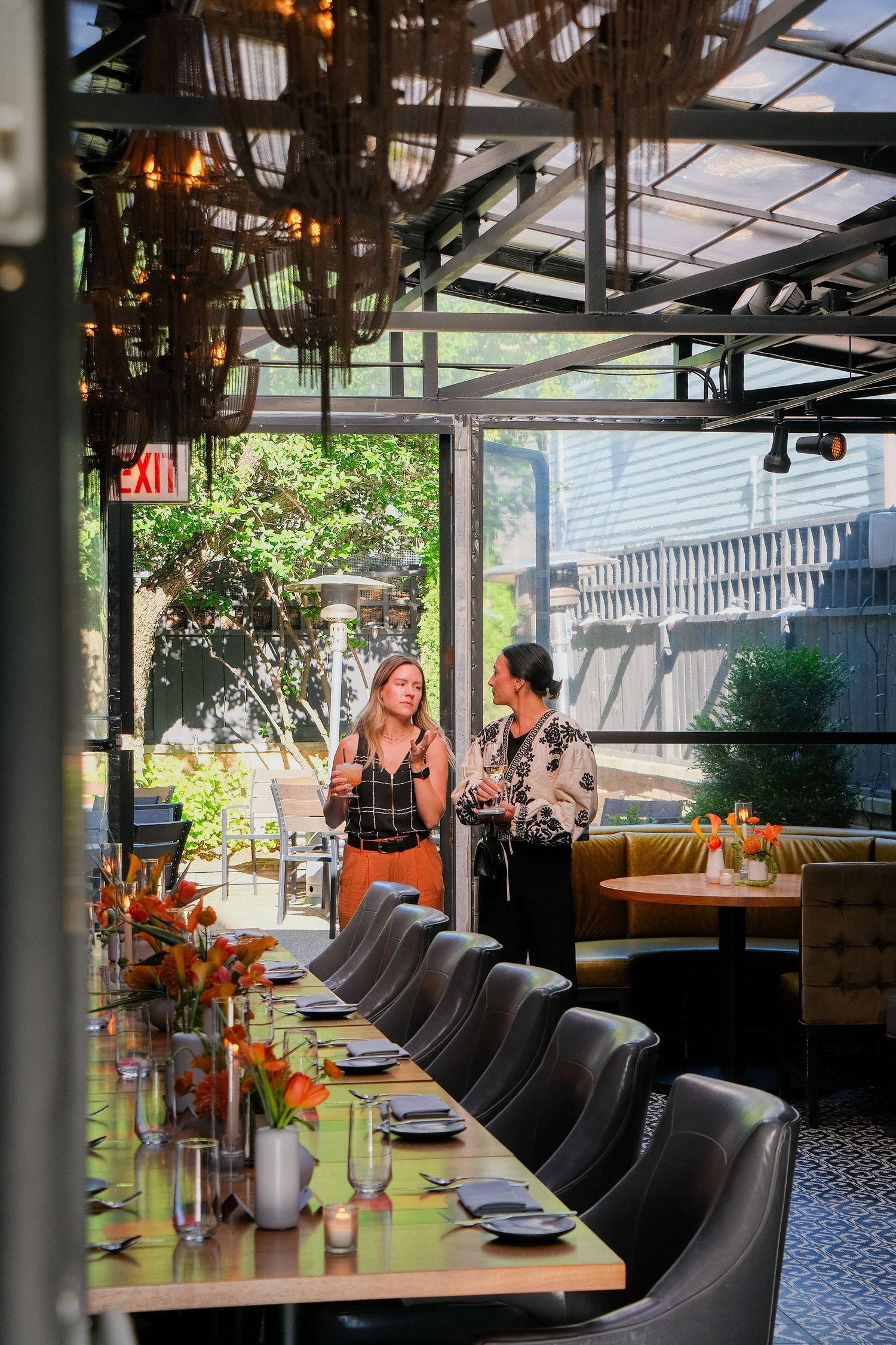 Two women conversing in an elegant restaurant with a long dining table, leather chairs, and orange floral centerpieces.
