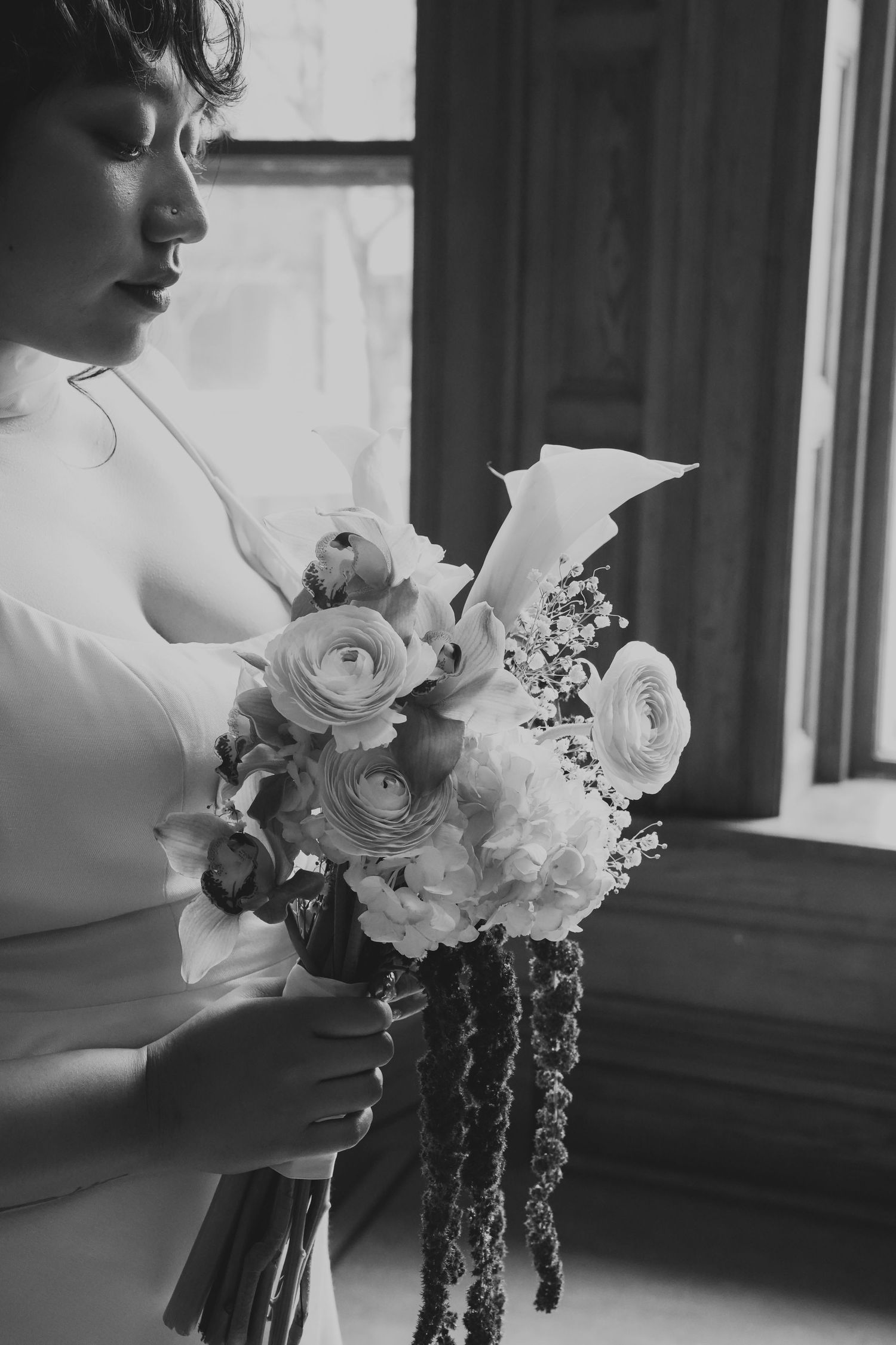 Black and white photo of a bride in white dress holding a bouquet of roses and calla lilies near a window.