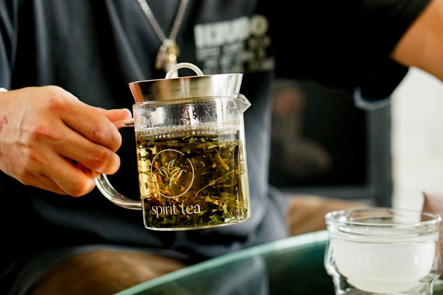 A hand pours golden liquid from a glass teapot into a small clear cup on a dark surface.