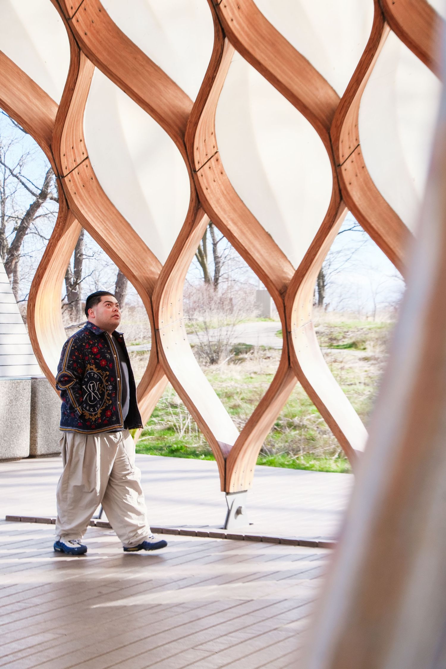 Man walking through an ornate wooden lattice pavilion with curved geometric patterns in a park setting.