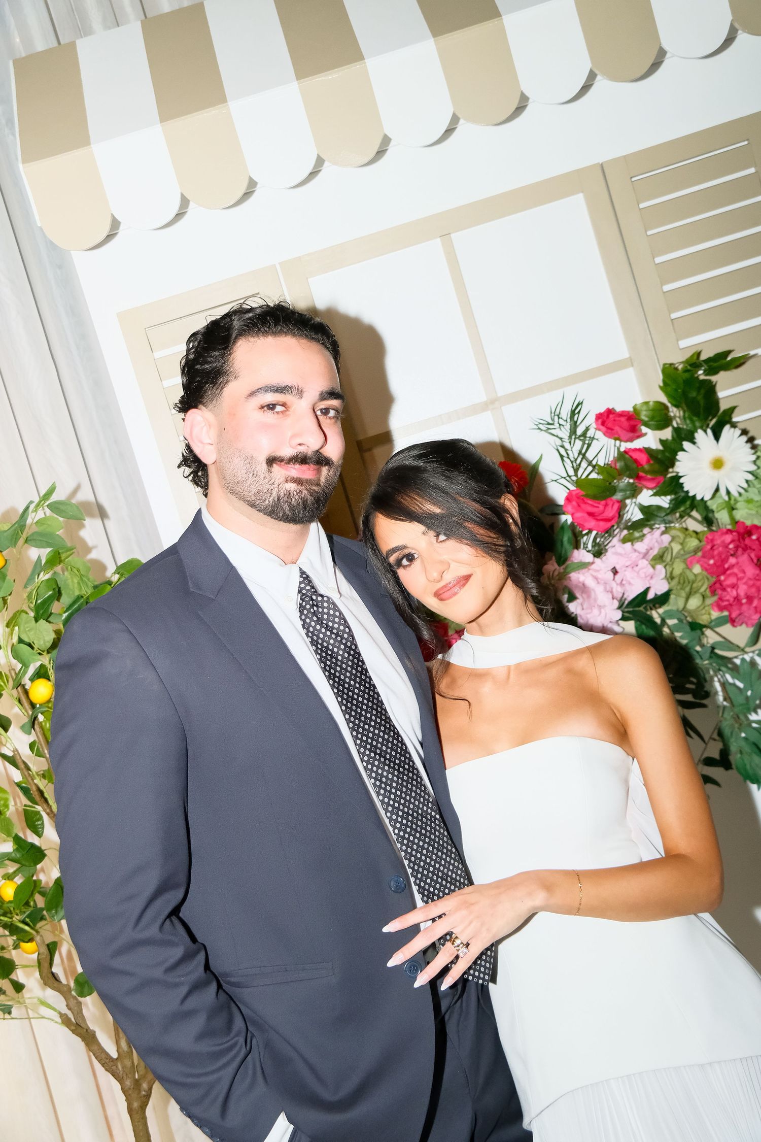 Bride and groom posing together near floral arrangements, man in gray suit and woman in white strapless dress.