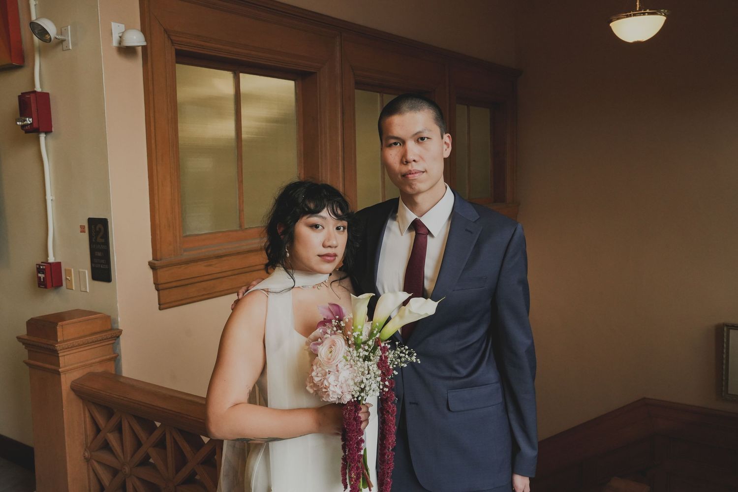 A bride and groom pose indoors on their wedding day, she in white holding calla lilies, he in a navy suit.