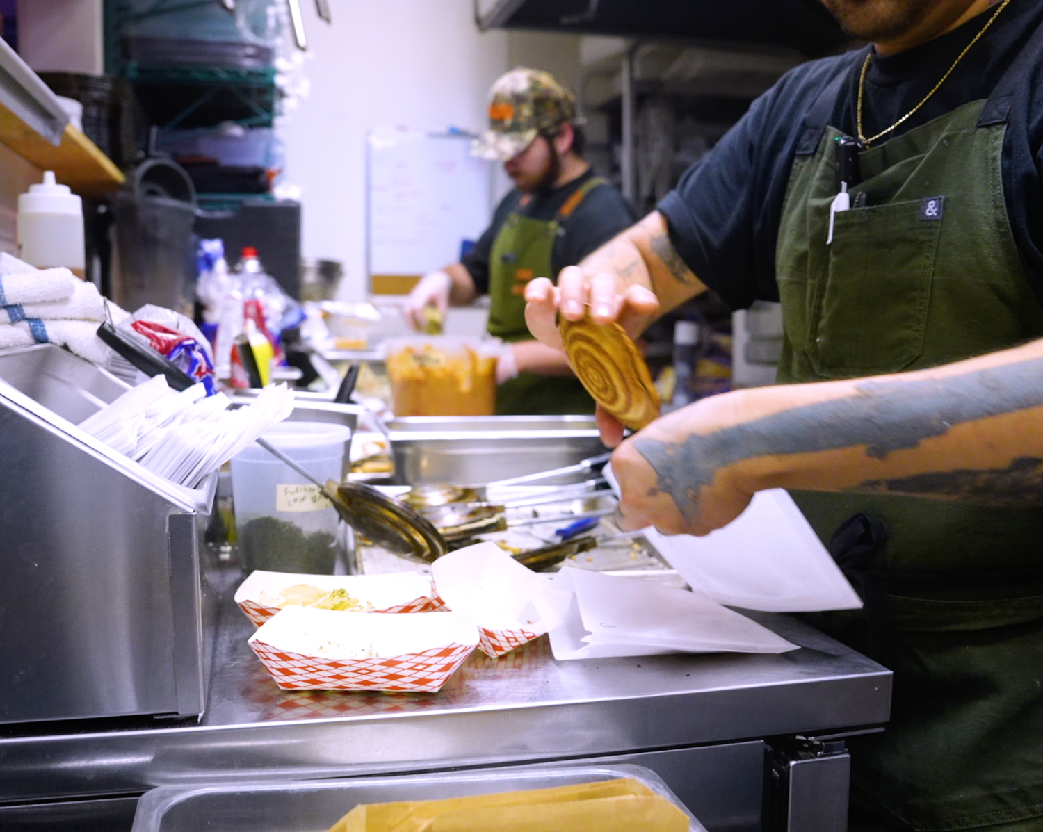 Two chefs in green aprons prepare food in a busy commercial kitchen, plating dishes at a stainless steel counter.