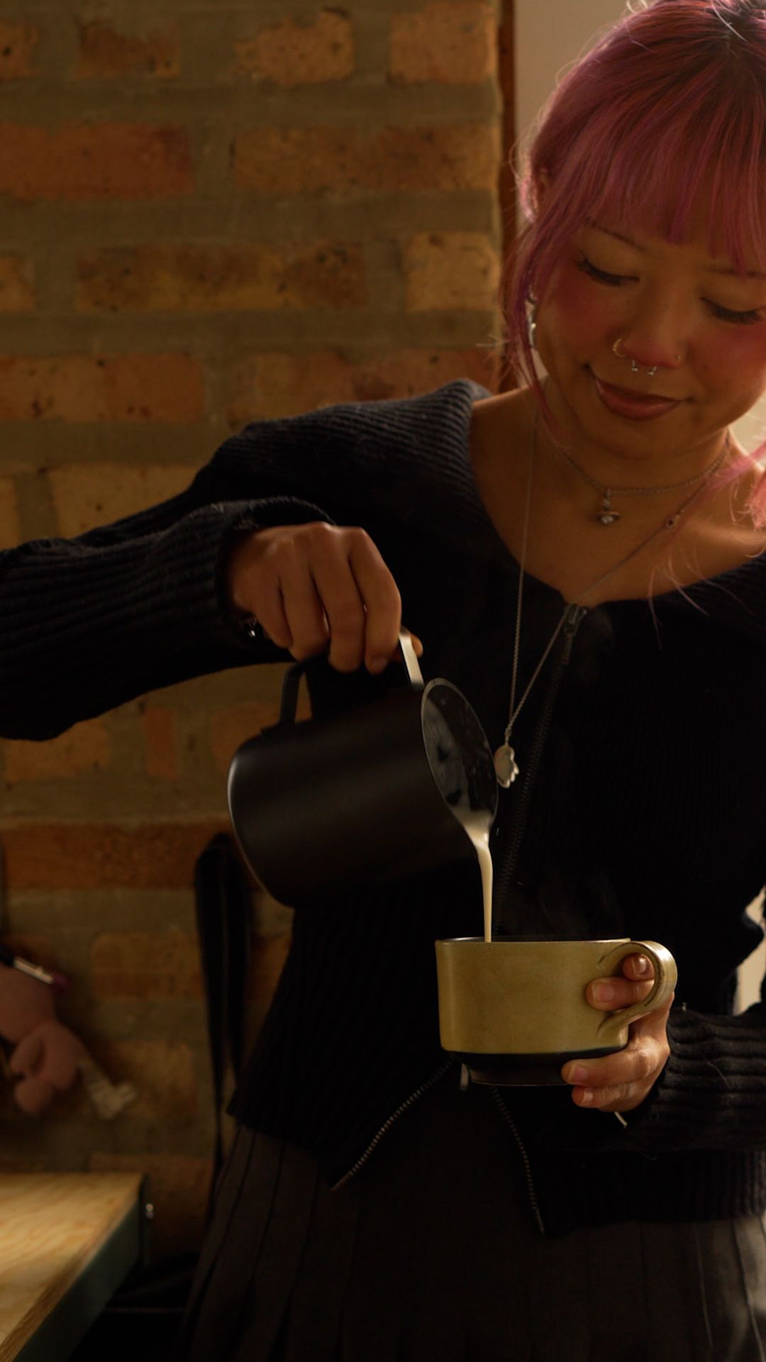 A person in black clothing stirs a warm beverage in a white cup against a brick wall background.