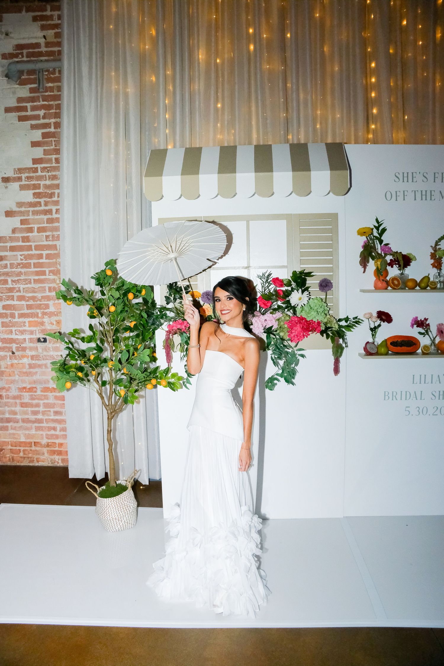 Bride in white ruffled gown holding parasol at bridal shower photo booth with floral decorations and string lights.