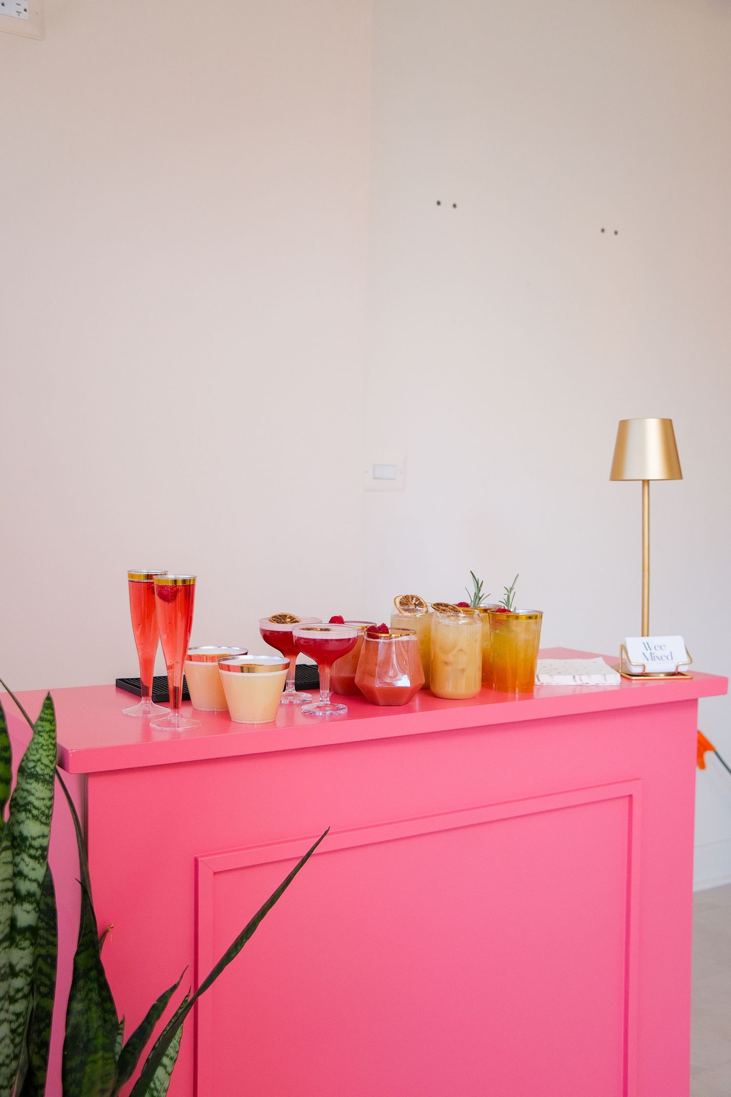 Pink bar counter with various colorful cocktails and drinks displayed against a white wall with green plants visible.