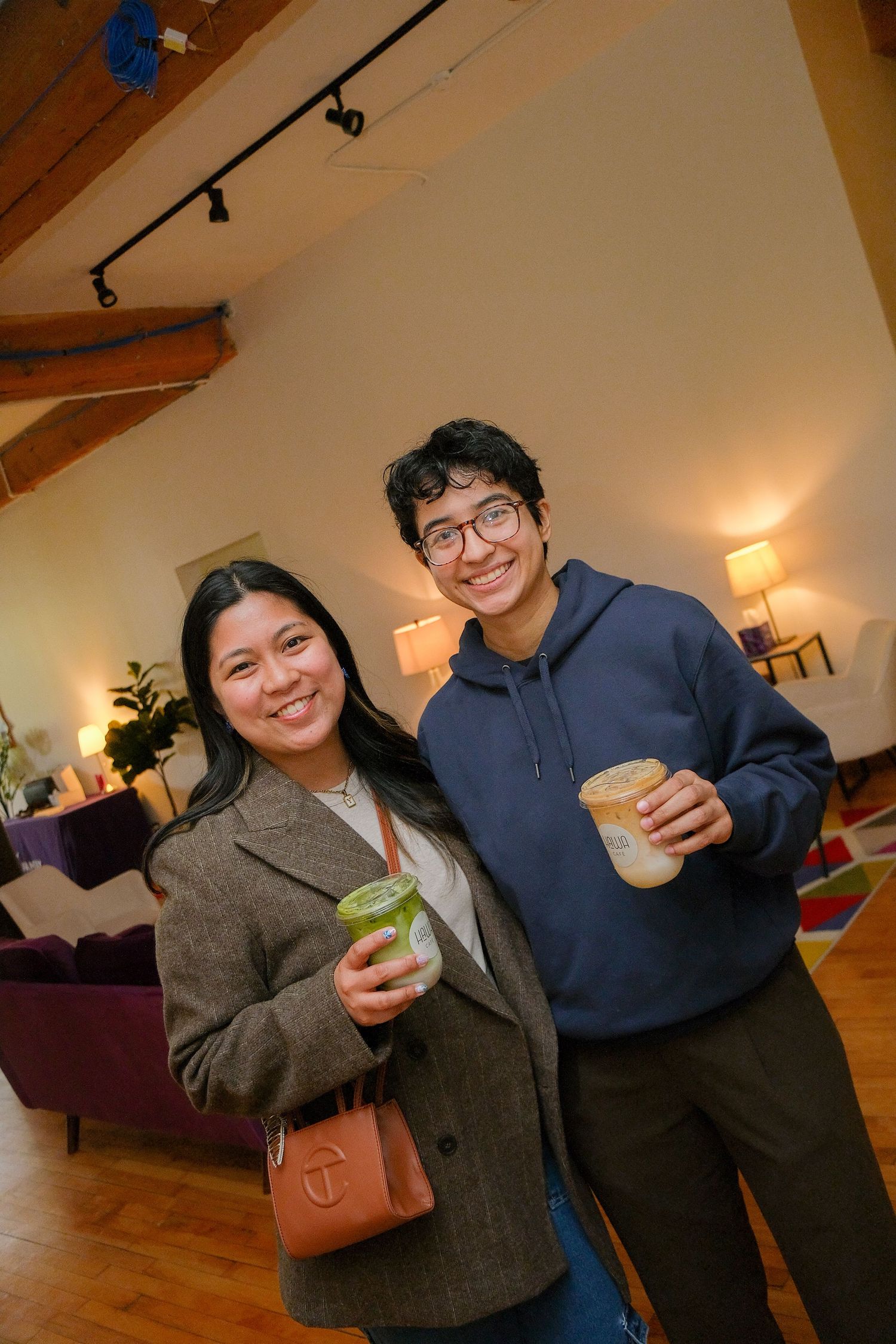 Two smiling friends holding coffee drinks pose together in a warmly lit modern event space.