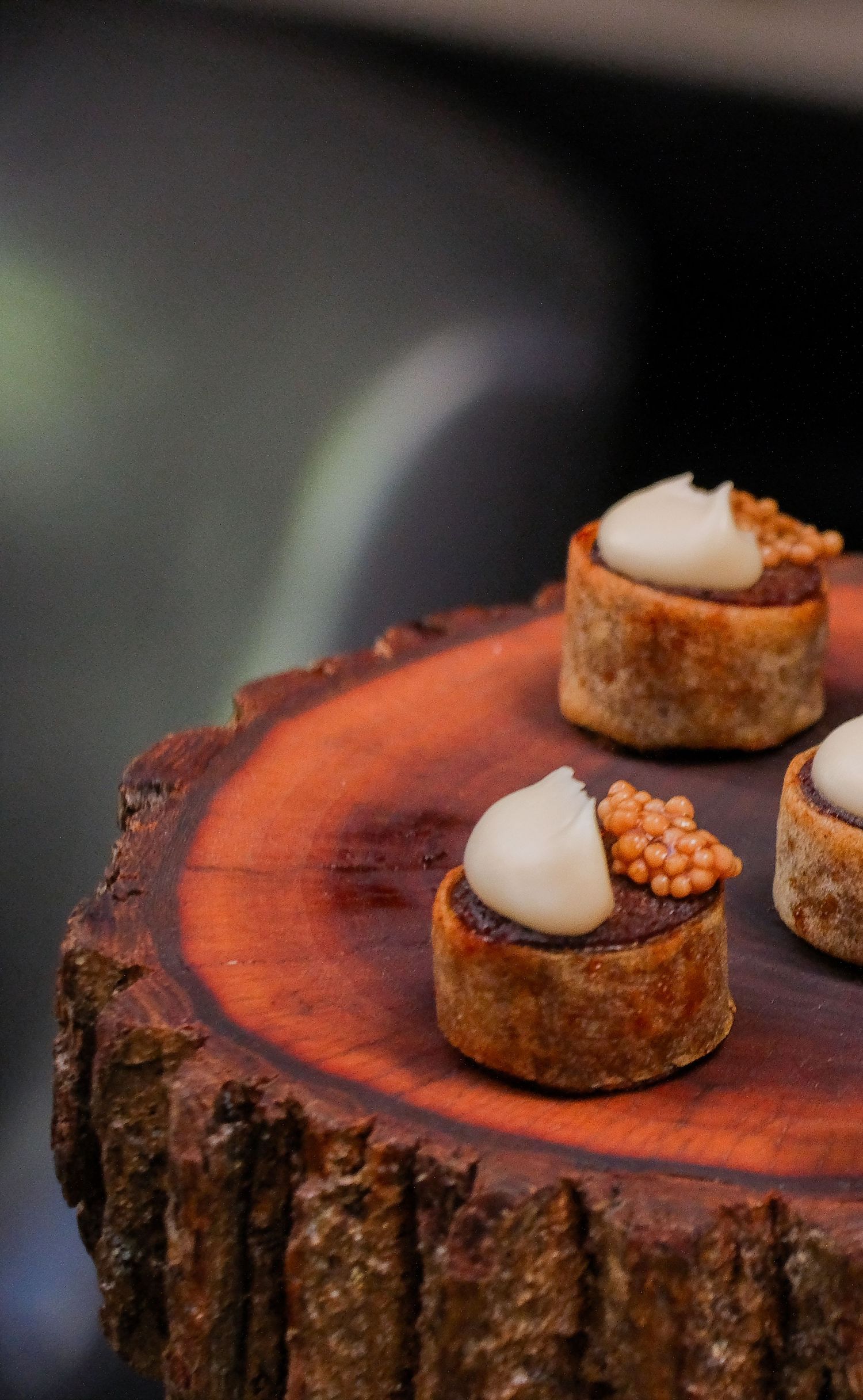 Elegant bite-sized pastry canapés topped with cream and orange caviar pearls, served on a rustic wooden slice.