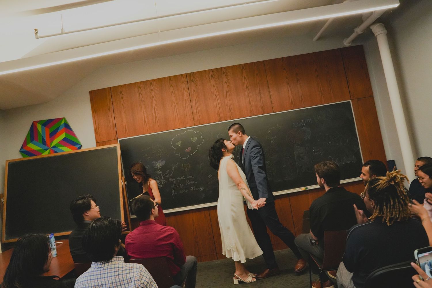 Couple sharing a romantic kiss in a university classroom with chalkboard backdrop during their wedding ceremony.
