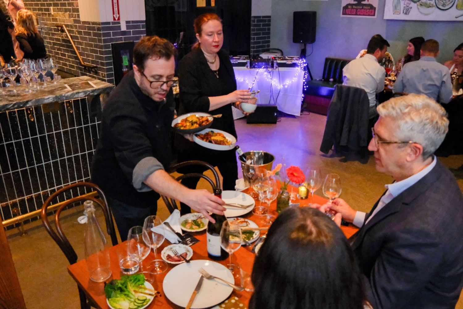 Chef serving food to guests at an elegant indoor dining event with waitstaff and attendees gathered around a table.