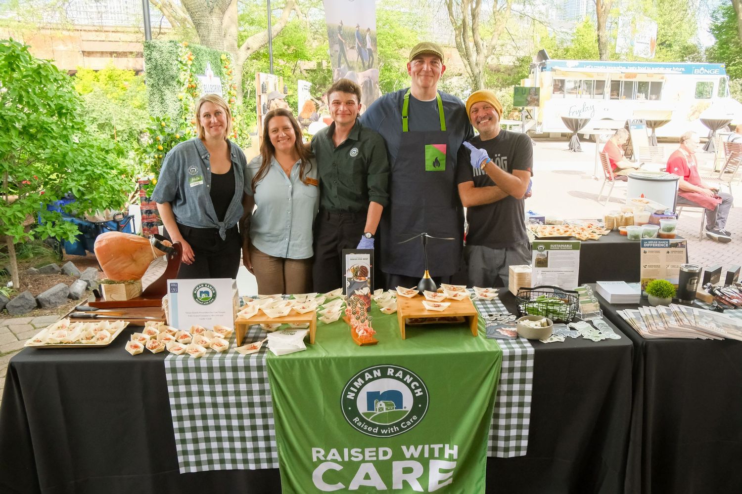 Five smiling vendors pose behind a table at an outdoor food market with a green checkered tablecloth and Raised With Care sign.