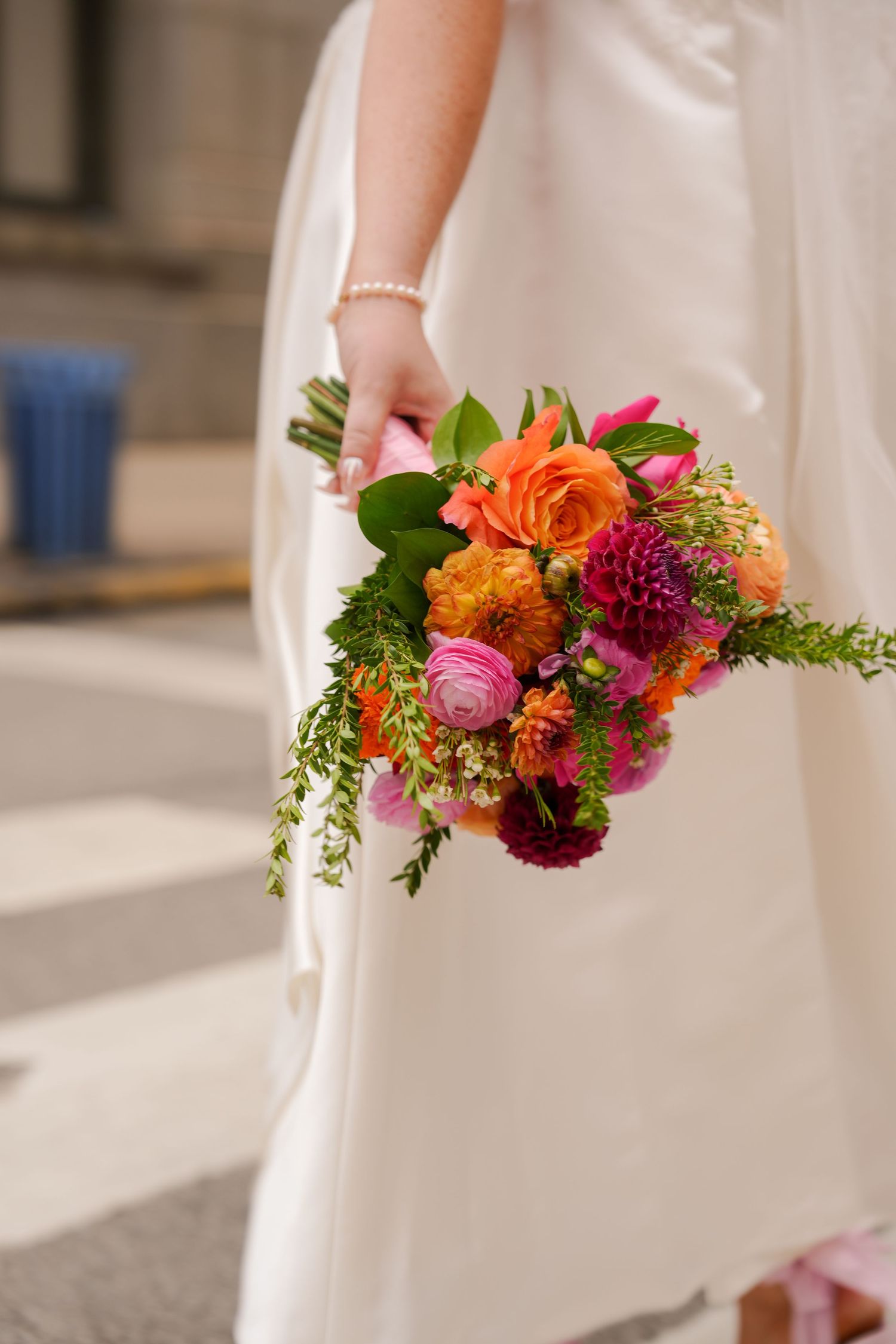 Bride in white gown holding vibrant bridal bouquet with orange roses, pink dahlias, and lush greenery on a city street.