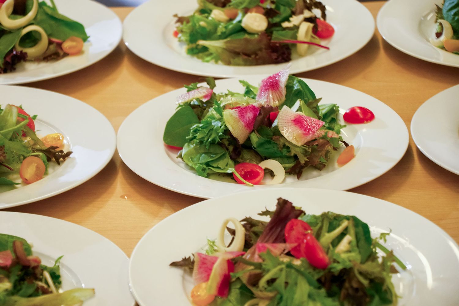 Fresh salad plates with mixed greens, garnishes and edible flowers arranged in a row on wooden table surface.